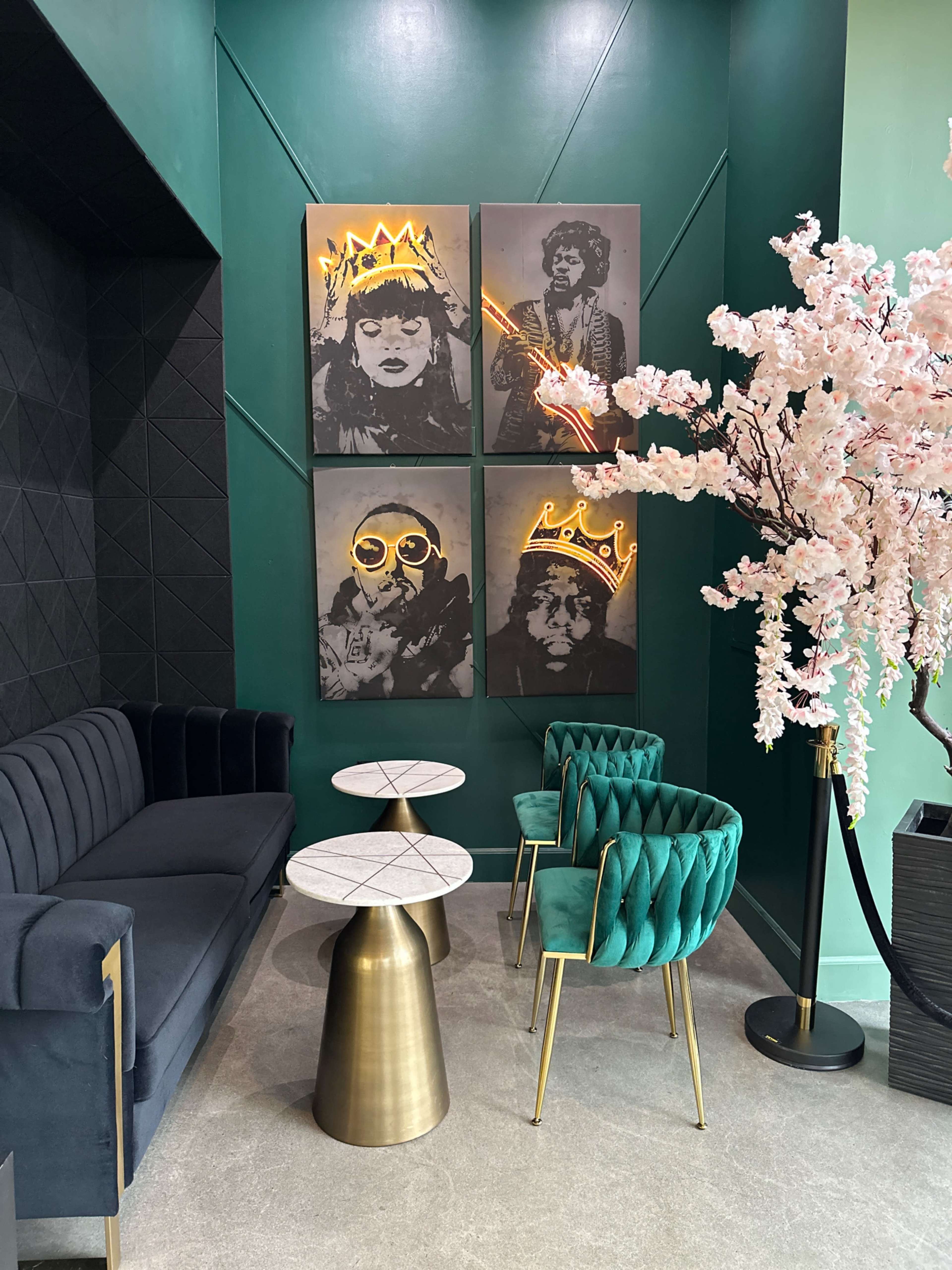 The image features a corner of a stylish café with two marble-top tables, green velvet chairs, a black sofa, and four framed portraits with neon accents on the walls, alongside a pink cherry blossom tree.
