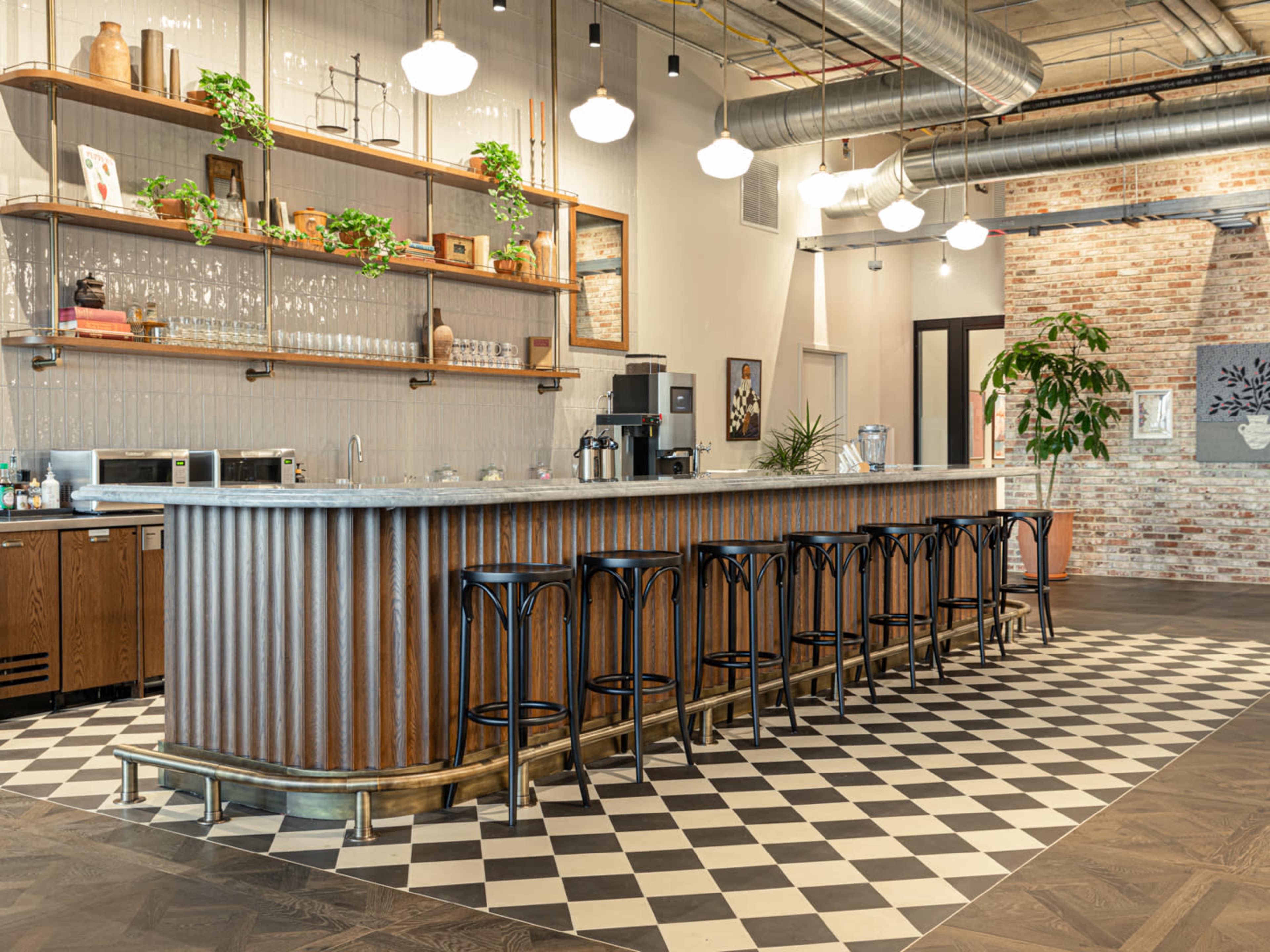 A modern bar area with a striped wooden counter, black stools, and a backdrop of shelves adorned with plants and glassware, all set against a checkered floor.