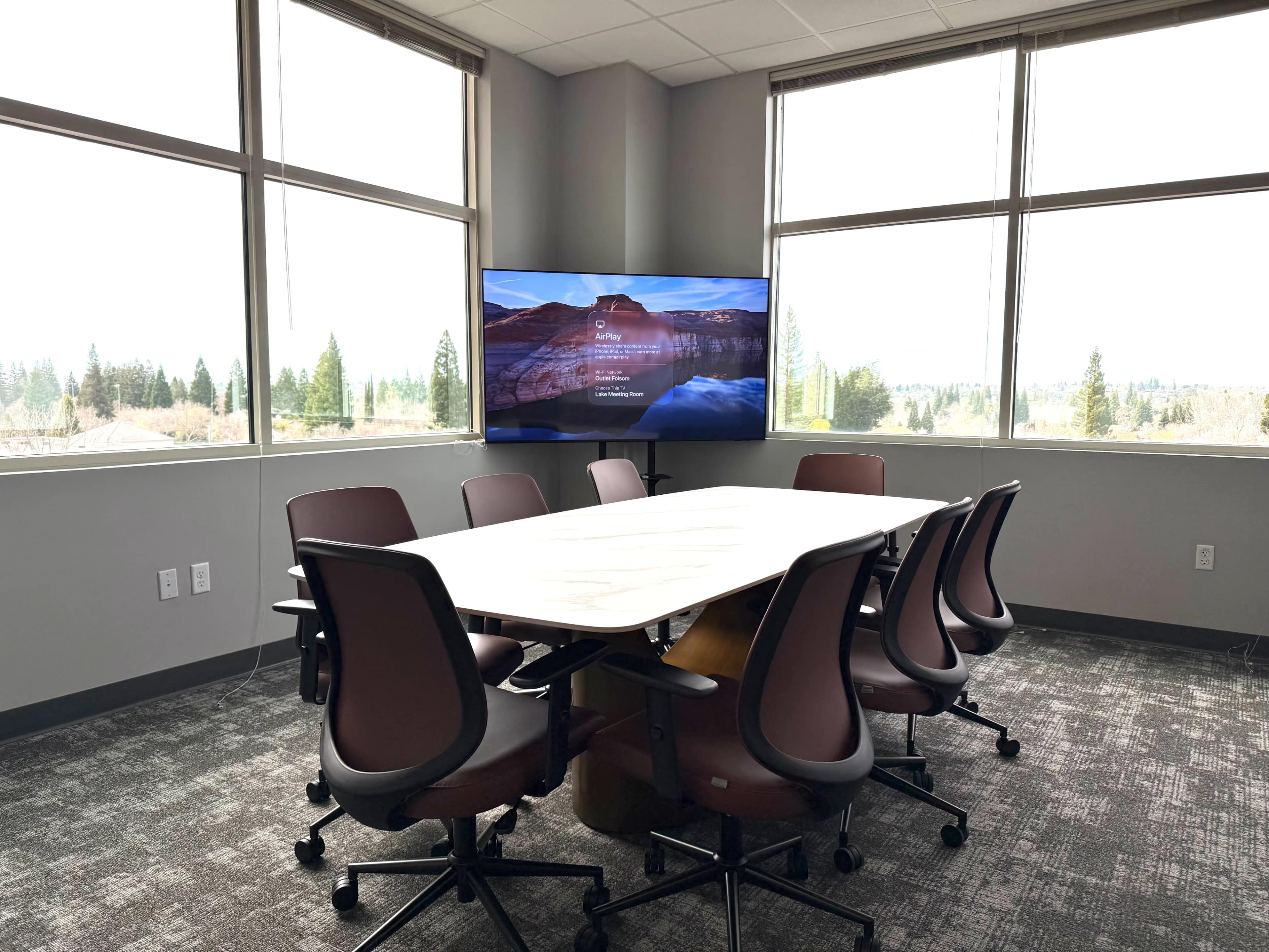 A conference room features a large table surrounded by eight rolling chairs, with a screen displaying content and large windows offering a view of trees outside.