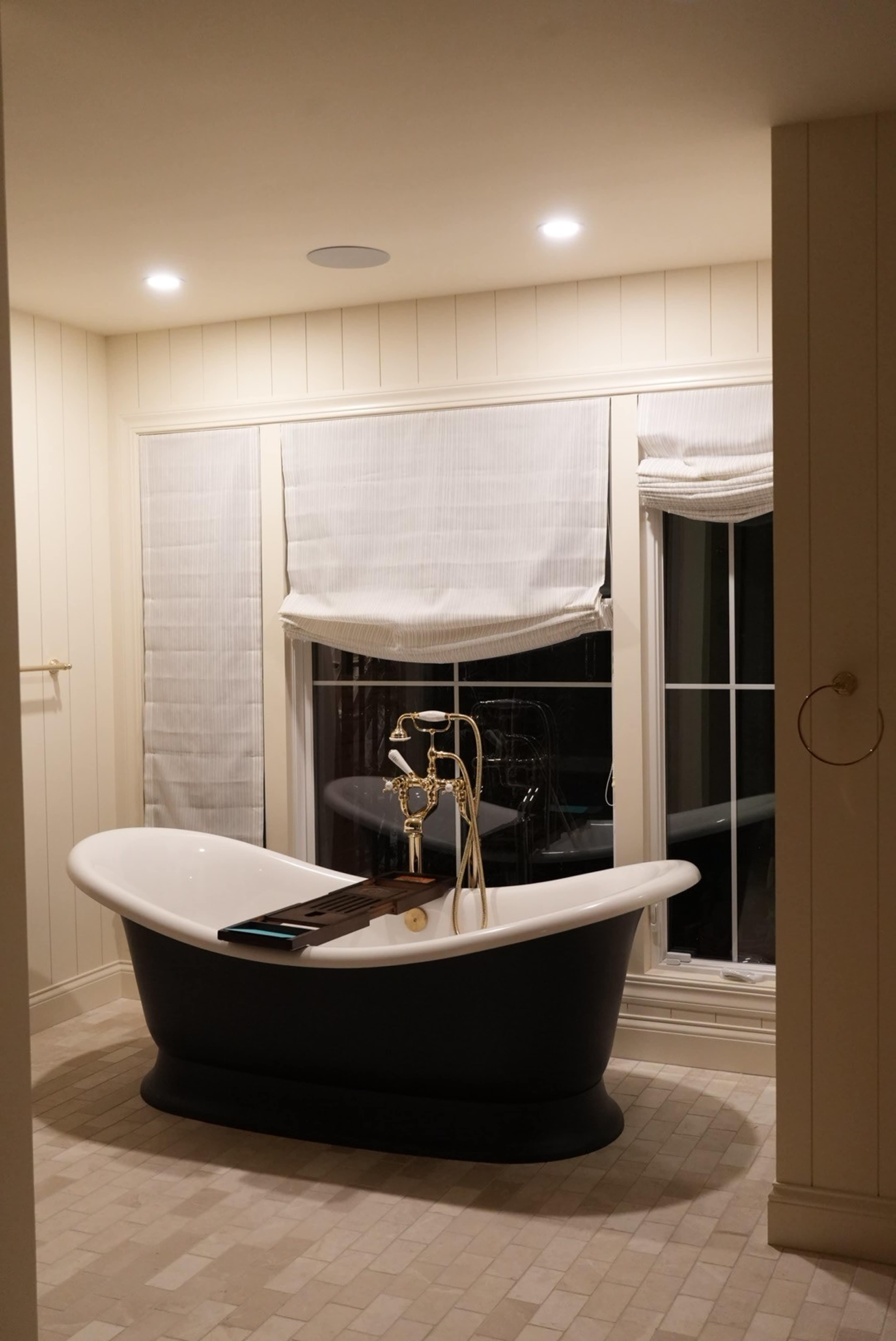 The image shows a modern bathroom featuring a freestanding bathtub near a large window adorned with shades.