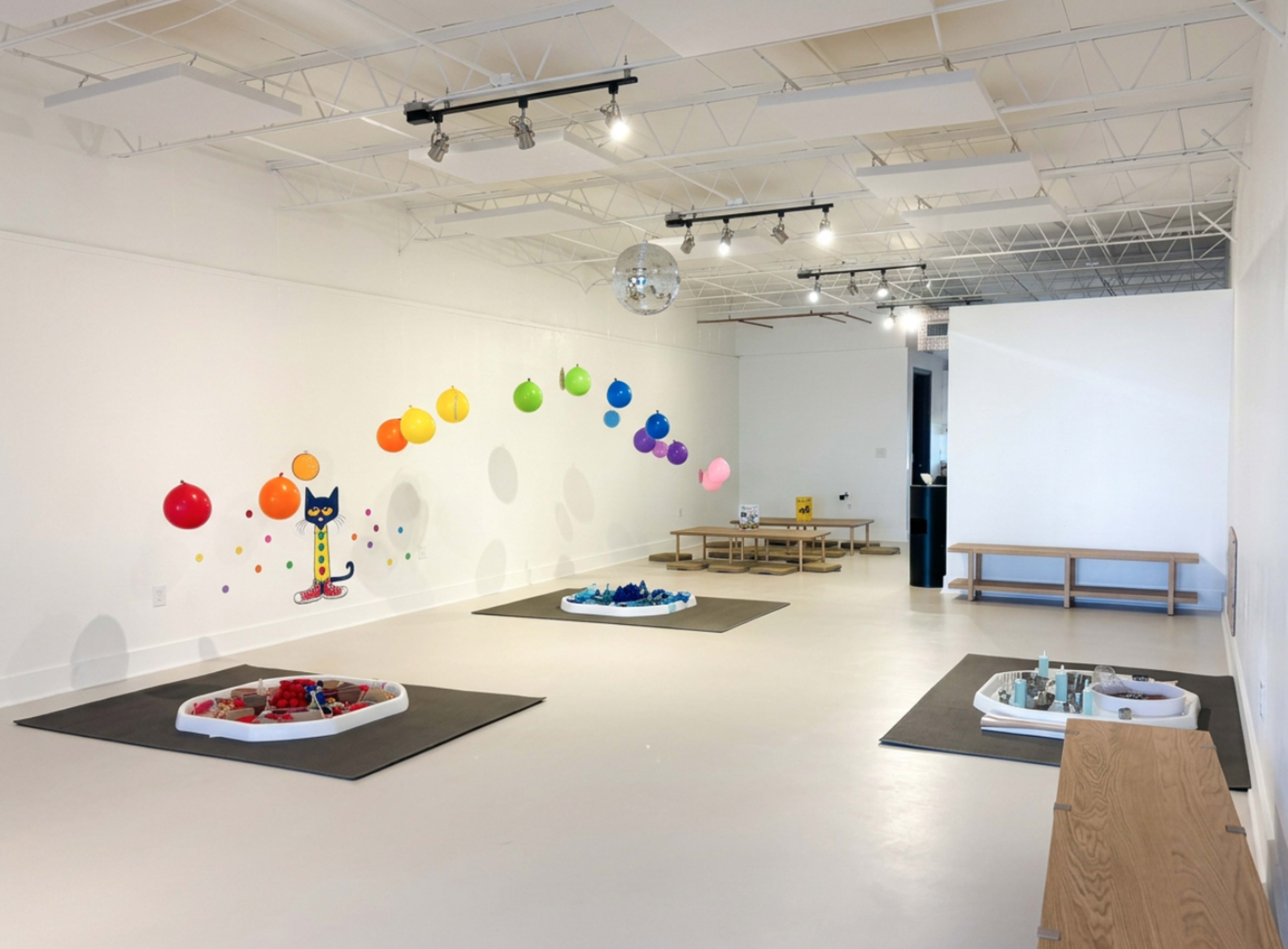 The image shows a spacious, brightly lit room featuring colorful balloons along the wall, several play areas with toys, and wooden benches.