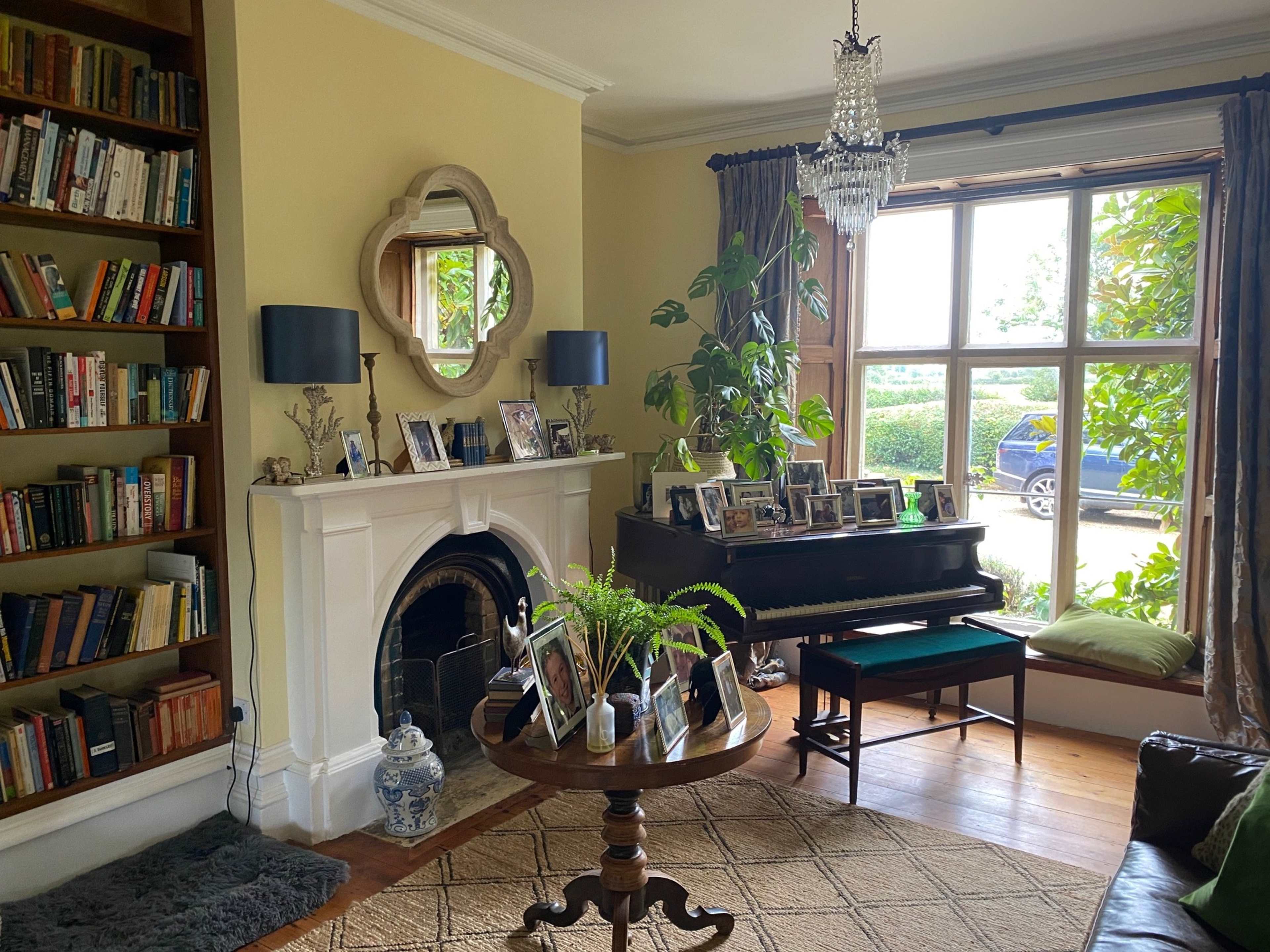 A living room features a piano, a mirror above the fireplace, shelves filled with books, and several framed photographs arranged on a table and the piano.