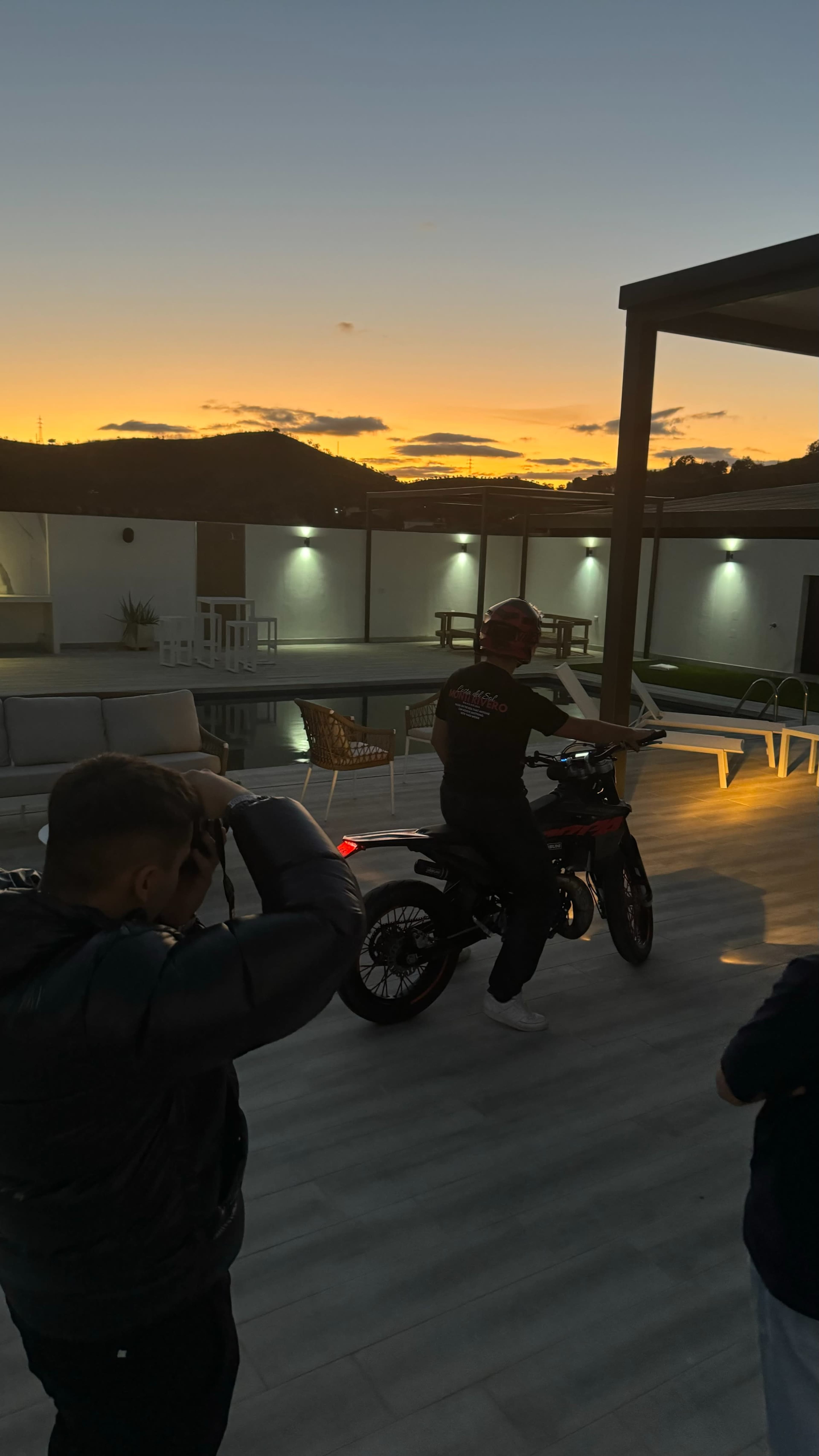 A motorcycle is positioned on a patio as two individuals take photographs against a colorful sunset backdrop.