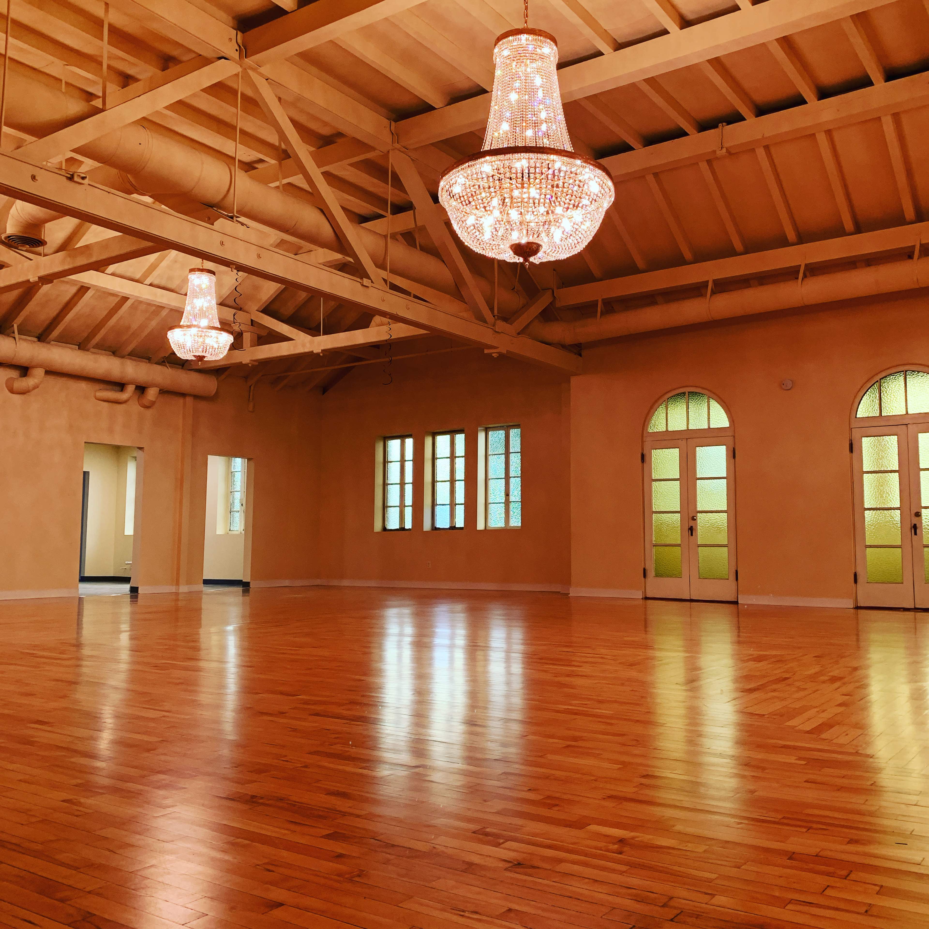 A spacious room with wooden floors, two chandeliers hanging from the ceiling, and several windows along the walls.