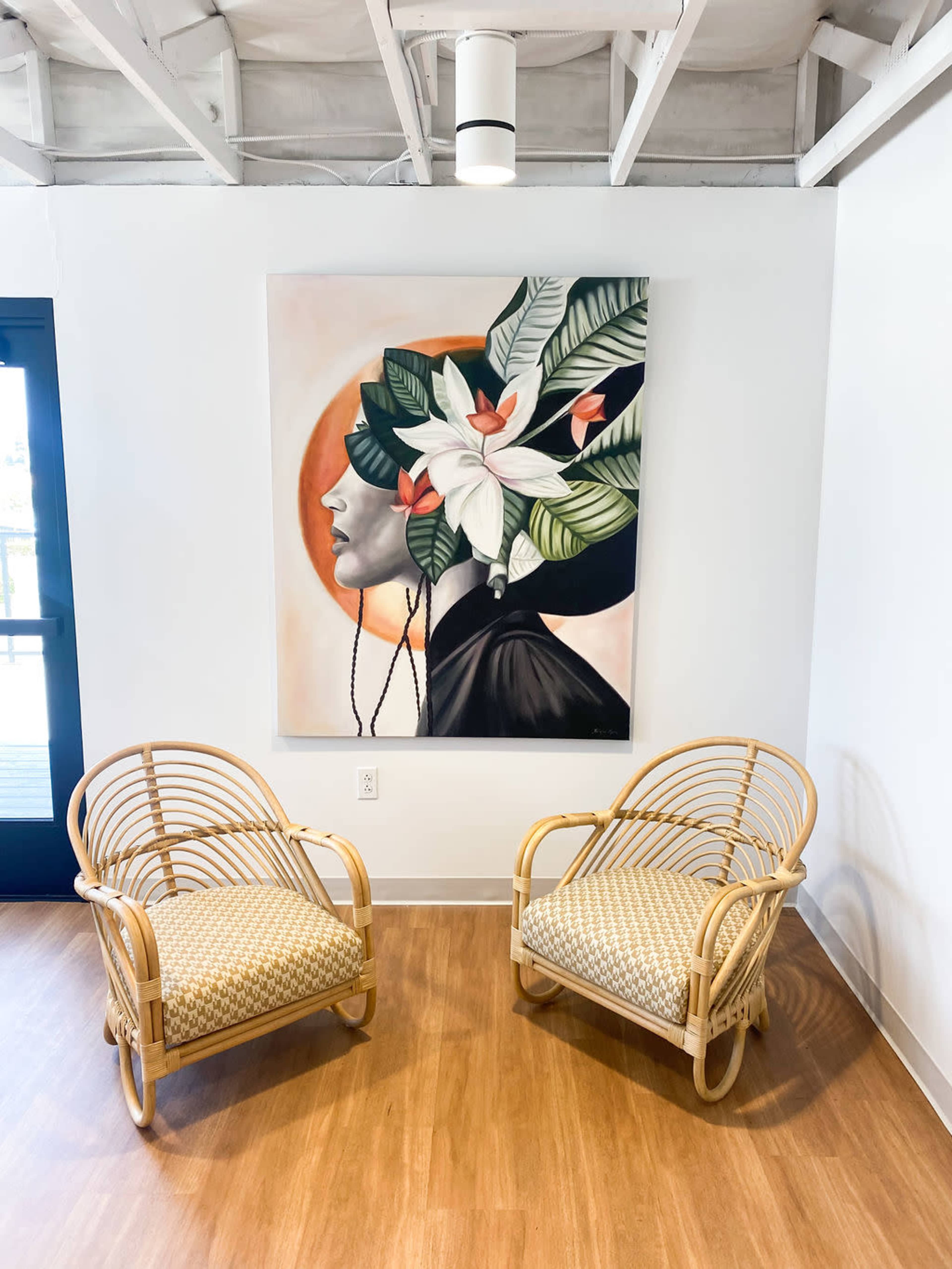 A pair of rattan chairs facing a large painting of a woman with floral elements on her head, positioned in a well-lit room with wooden flooring.