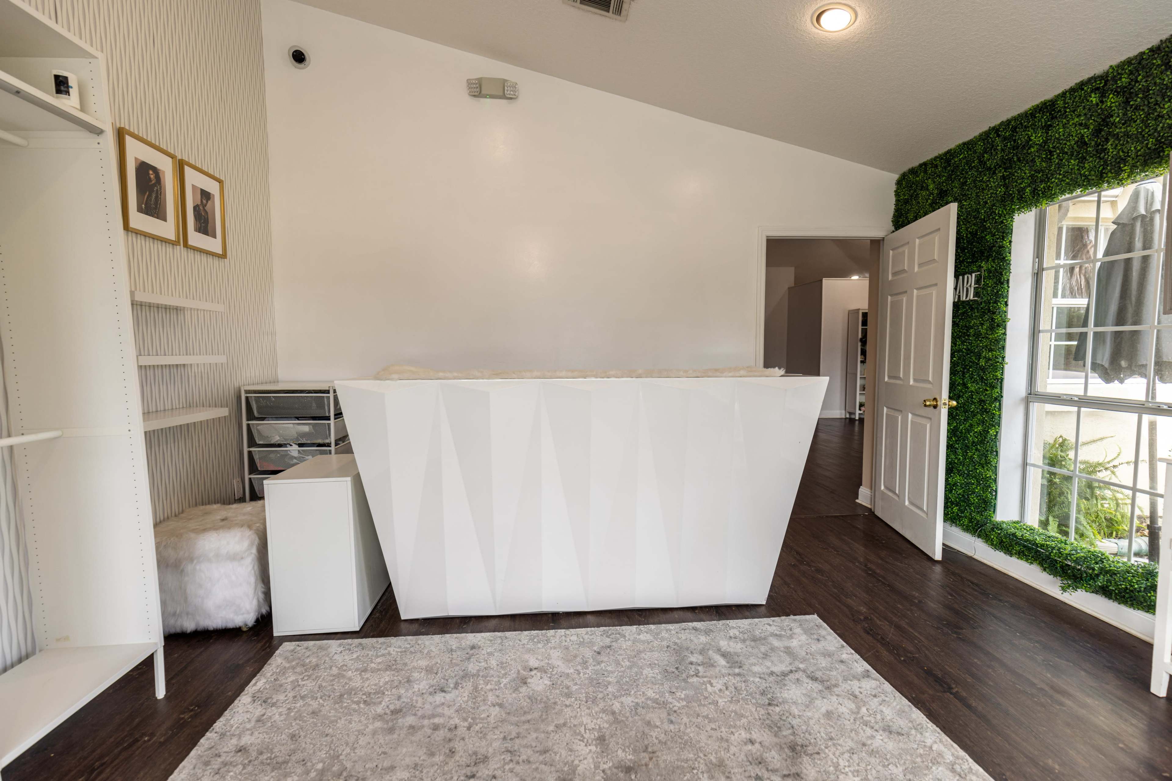 A modern reception area featuring a white geometric desk, a shelving unit, and a wall adorned with green foliage, with a door leading to another room in the background.