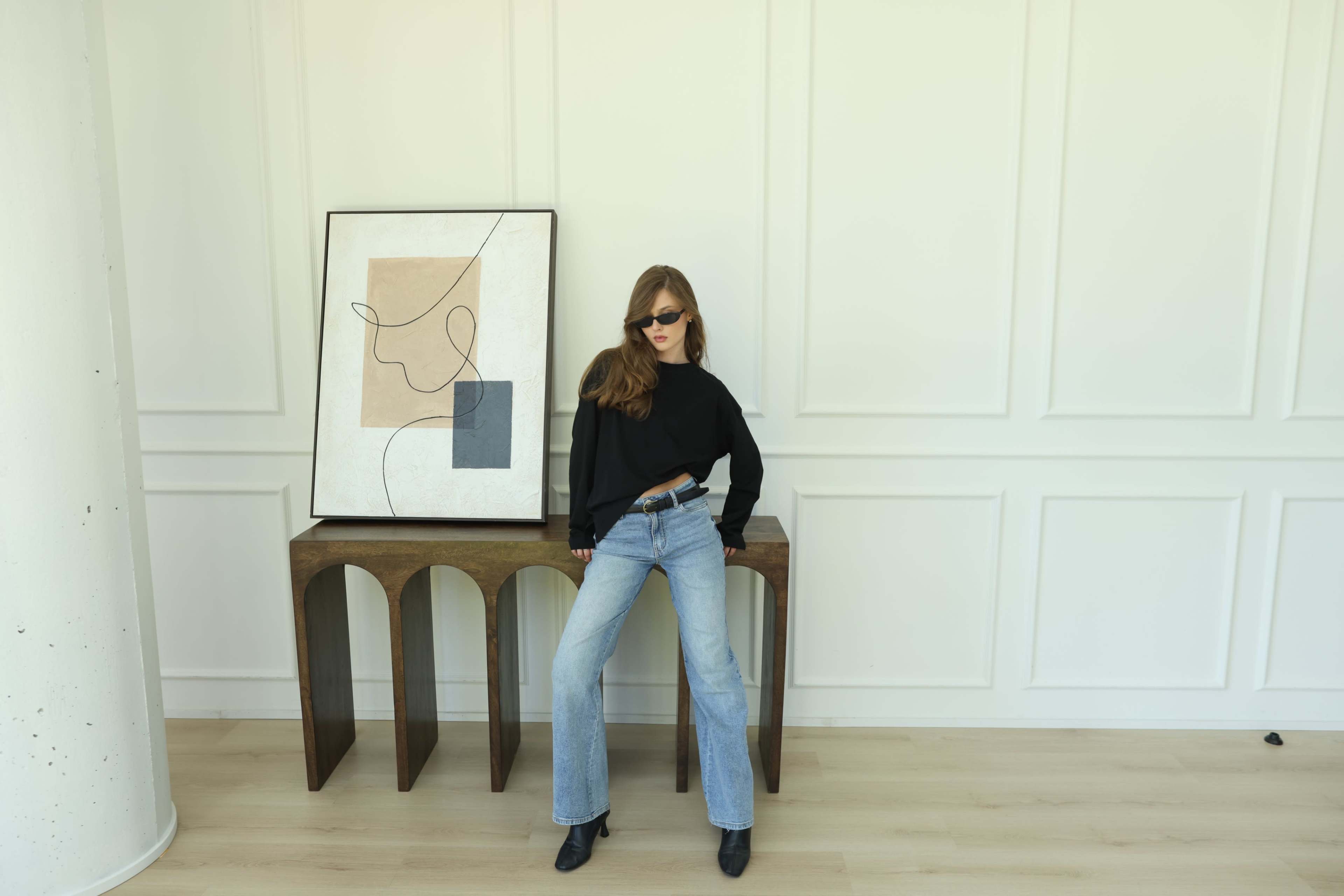 A woman wearing sunglasses stands confidently beside a modern artwork on a wooden console table in a minimalist interior.
