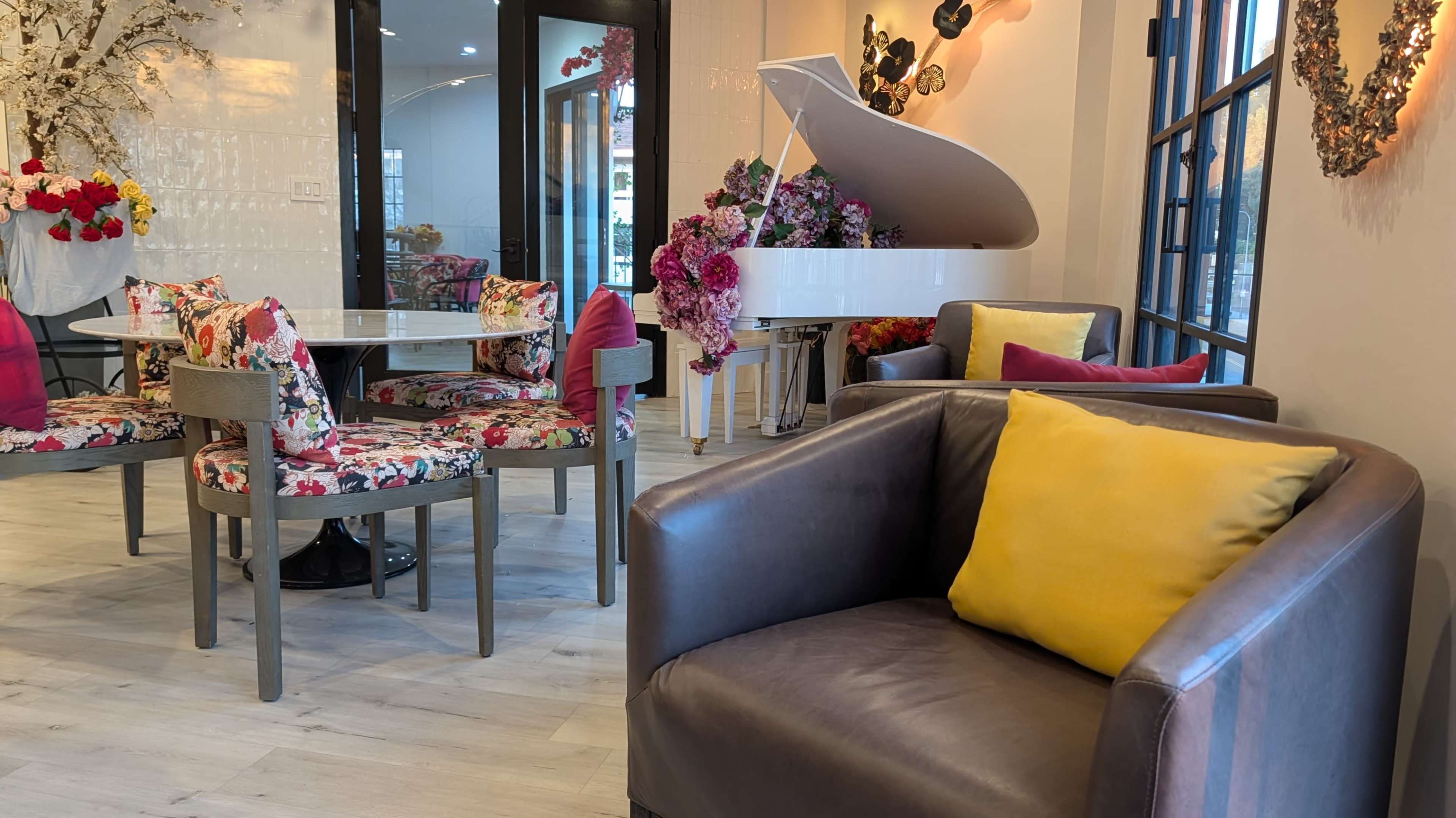 The image shows a stylish cafe interior featuring a white piano adorned with flowers, a round dining table surrounded by floral-patterned chairs, and modern seating arrangements with colorful cushions.