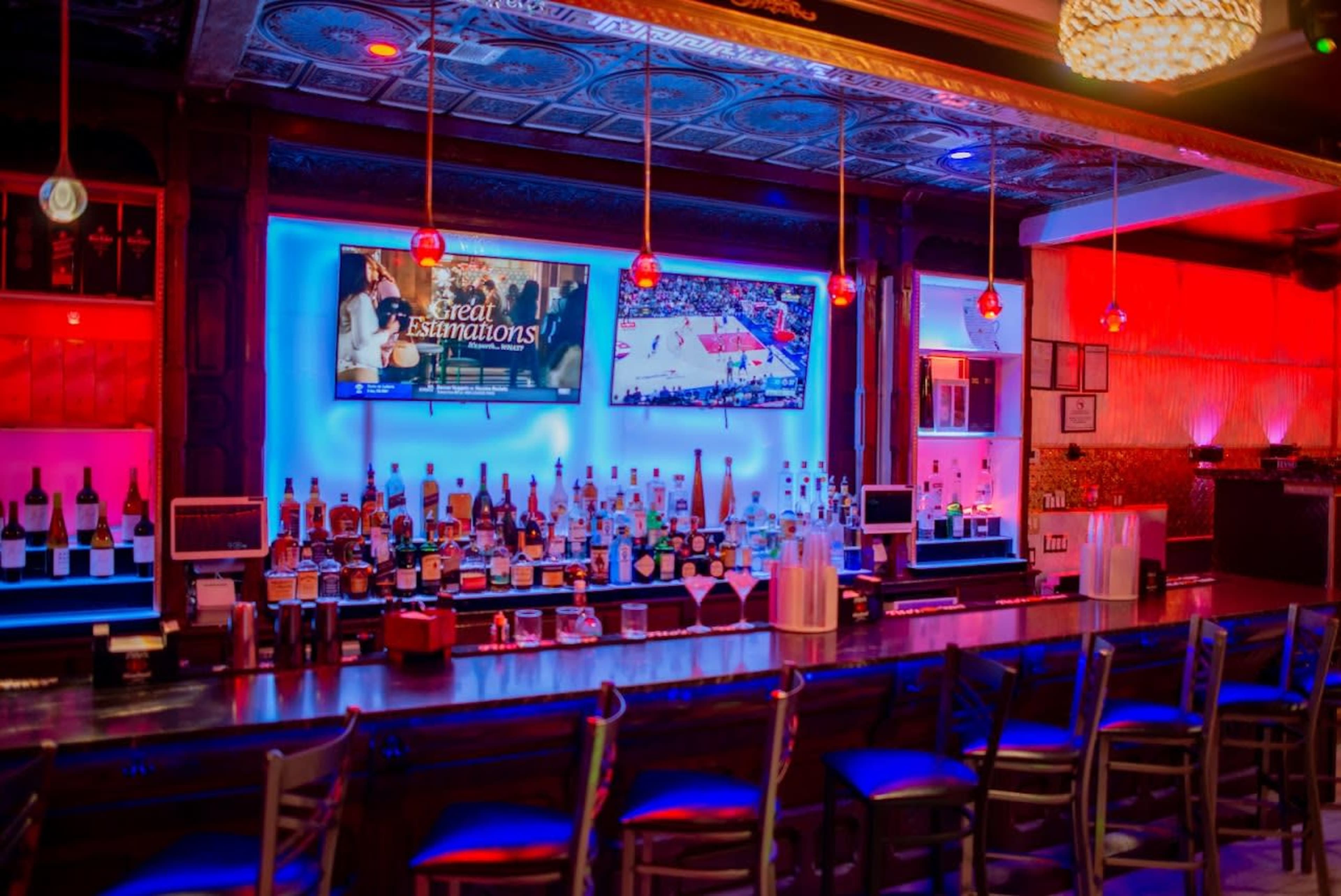 The image shows a brightly lit bar with a variety of alcoholic beverages displayed on shelves, two television screens showing sports, and colorful ambient lighting.