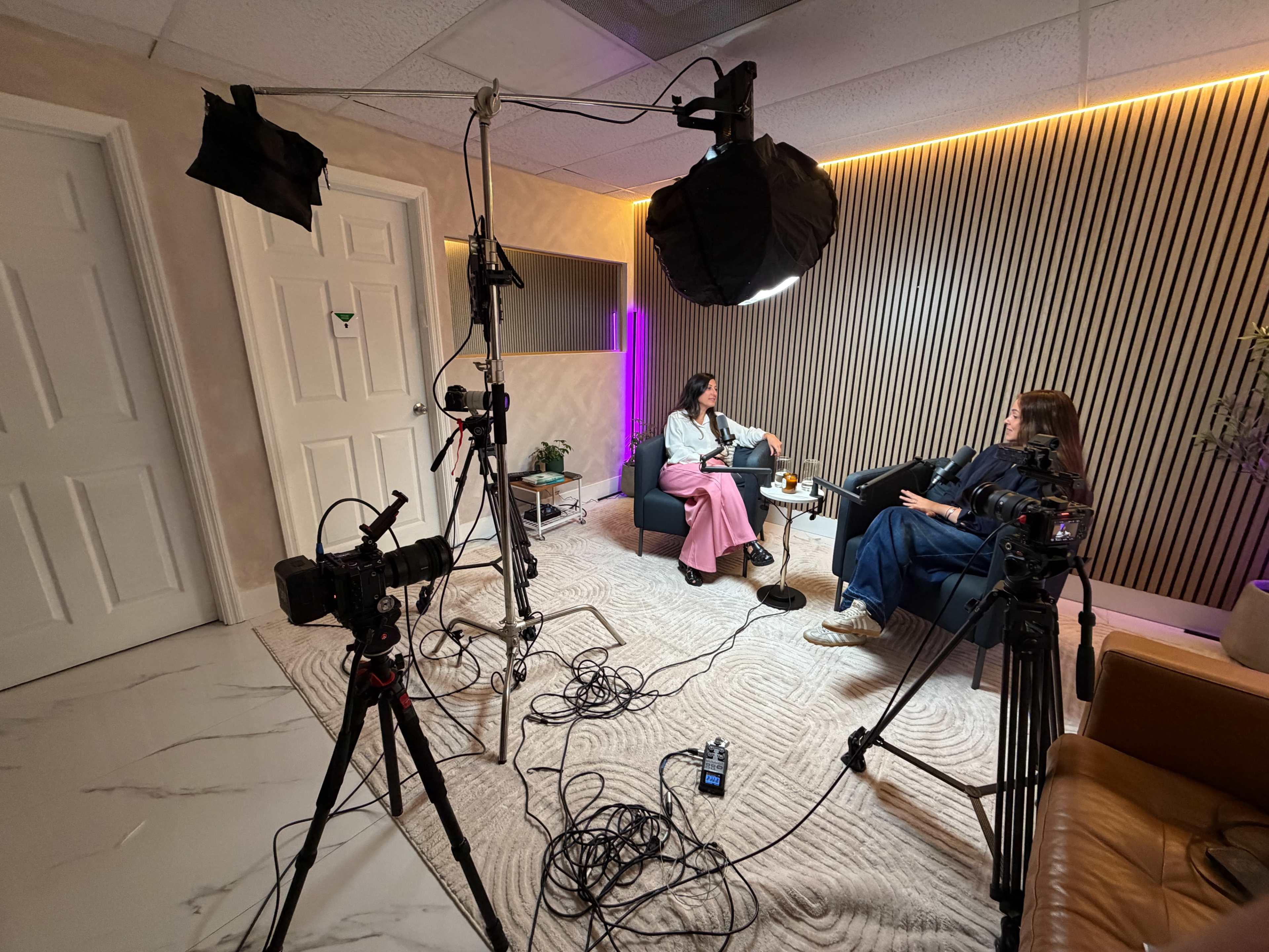 The image shows a well-lit studio setup for an interview, featuring two people seated on a couch, surrounded by cameras and lighting equipment.