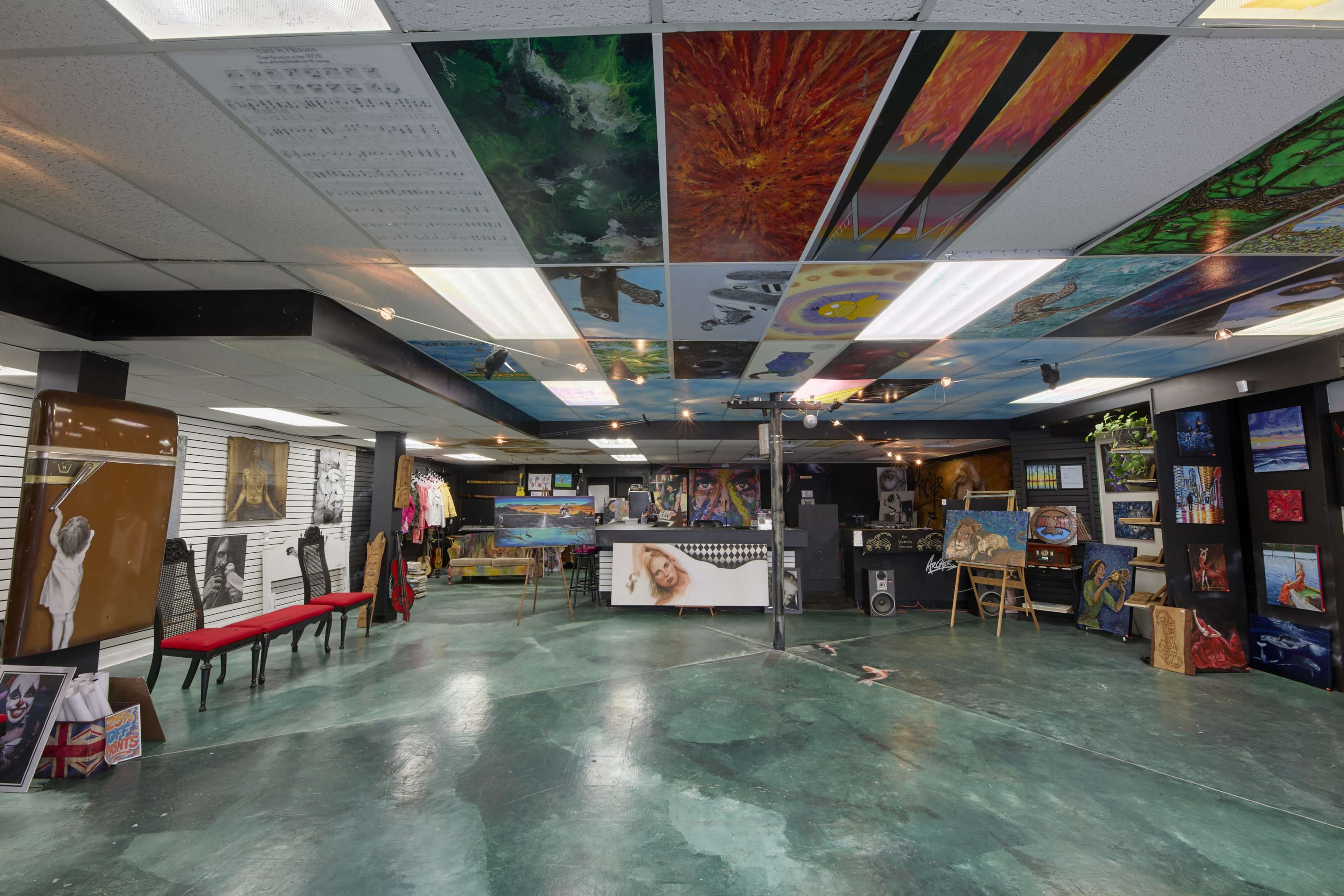 The image shows an art gallery with a colorful ceiling painted with various artworks, displaying paintings and clothing on racks in a spacious area.