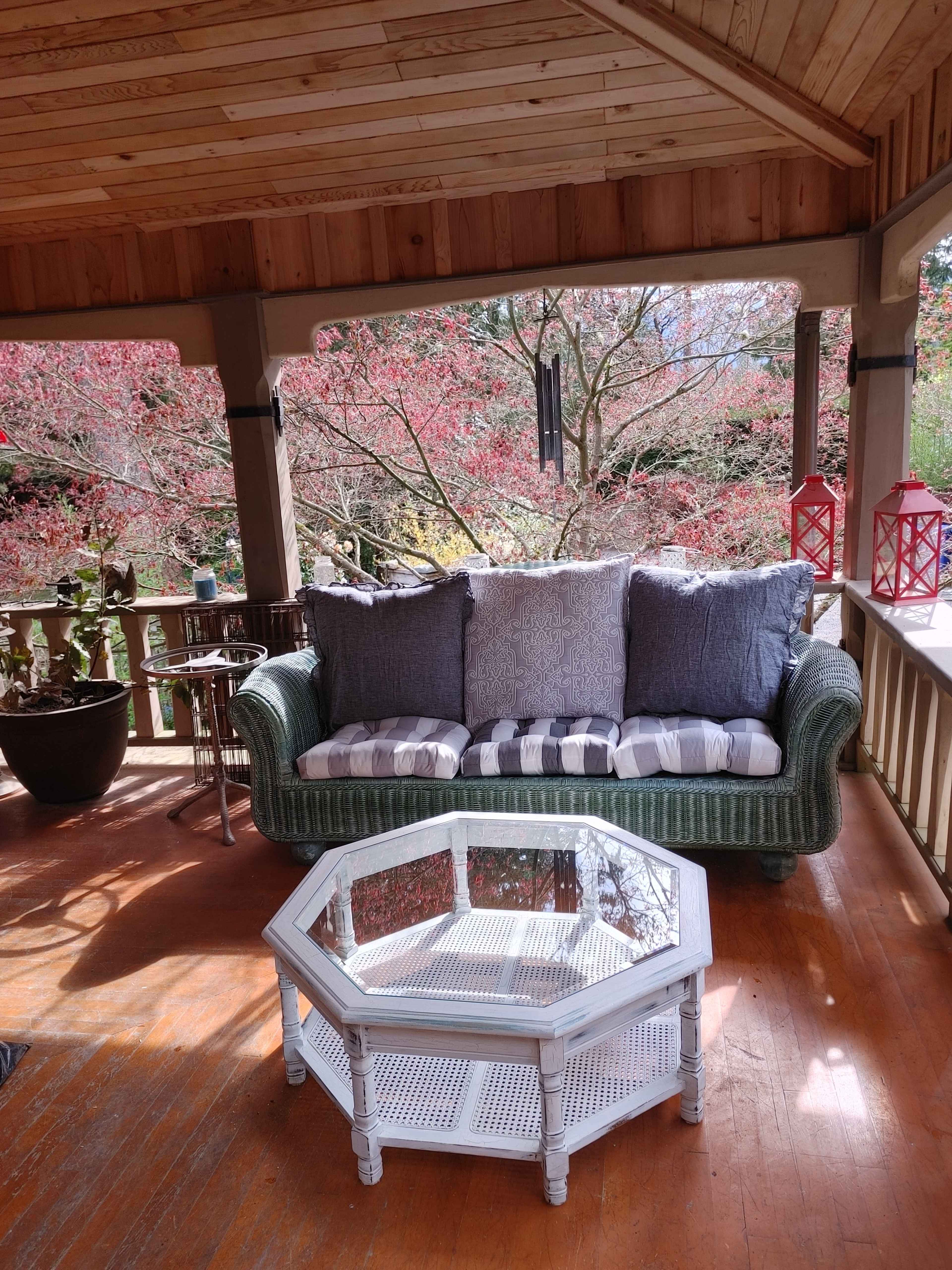 A wicker sofa with cushions is positioned beside an octagonal white coffee table on a porch, overlooking trees with pink foliage.