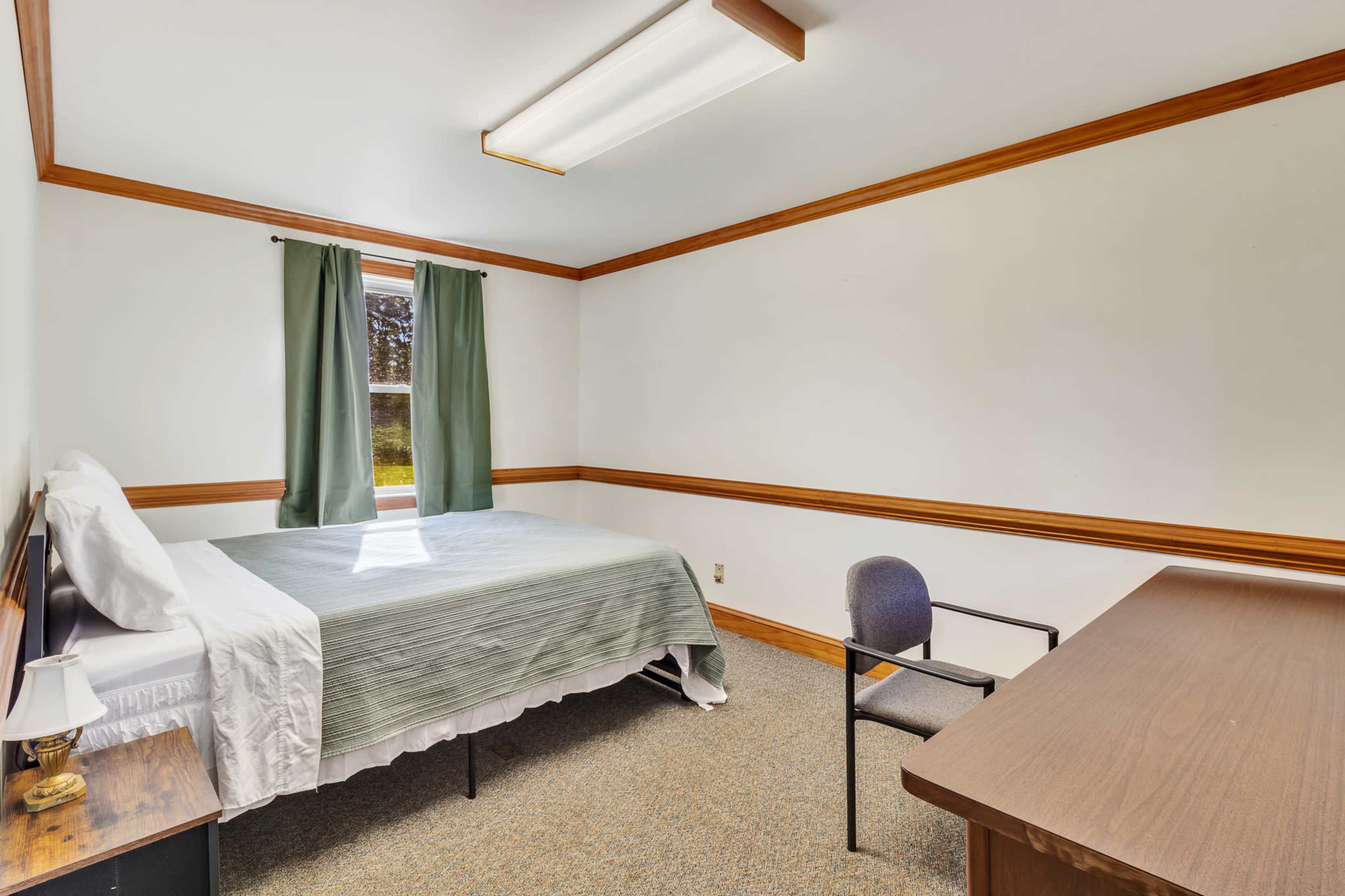The image shows a simple bedroom with a bed, a chair, a desk, and a window dressed with green curtains.