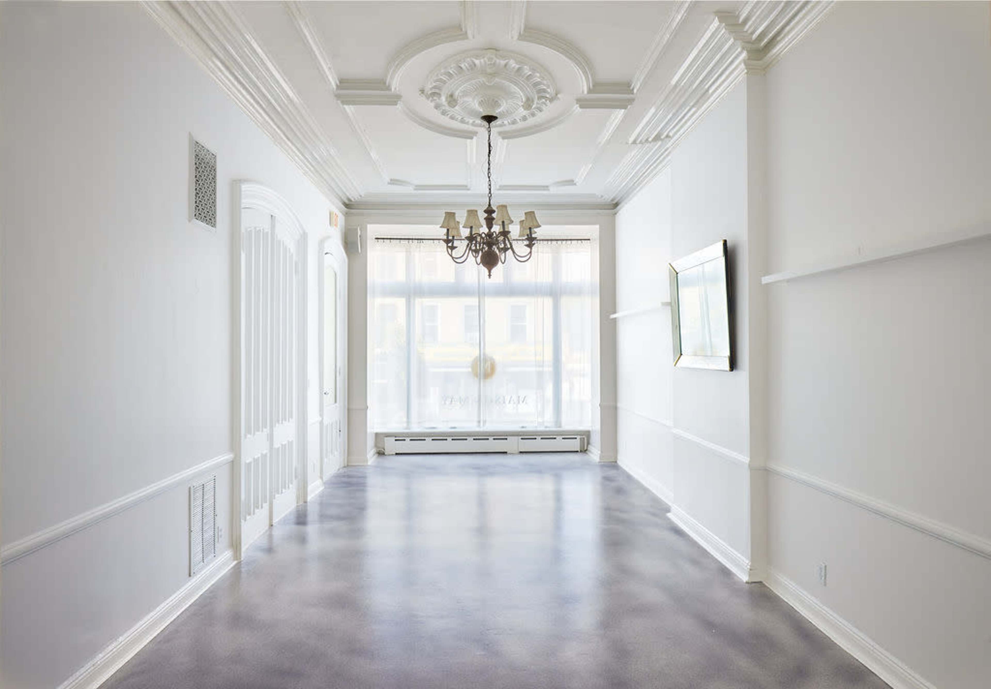 The image shows a spacious, well-lit interior room with a chandelier, large windows, and decorative molding on the ceiling and walls.