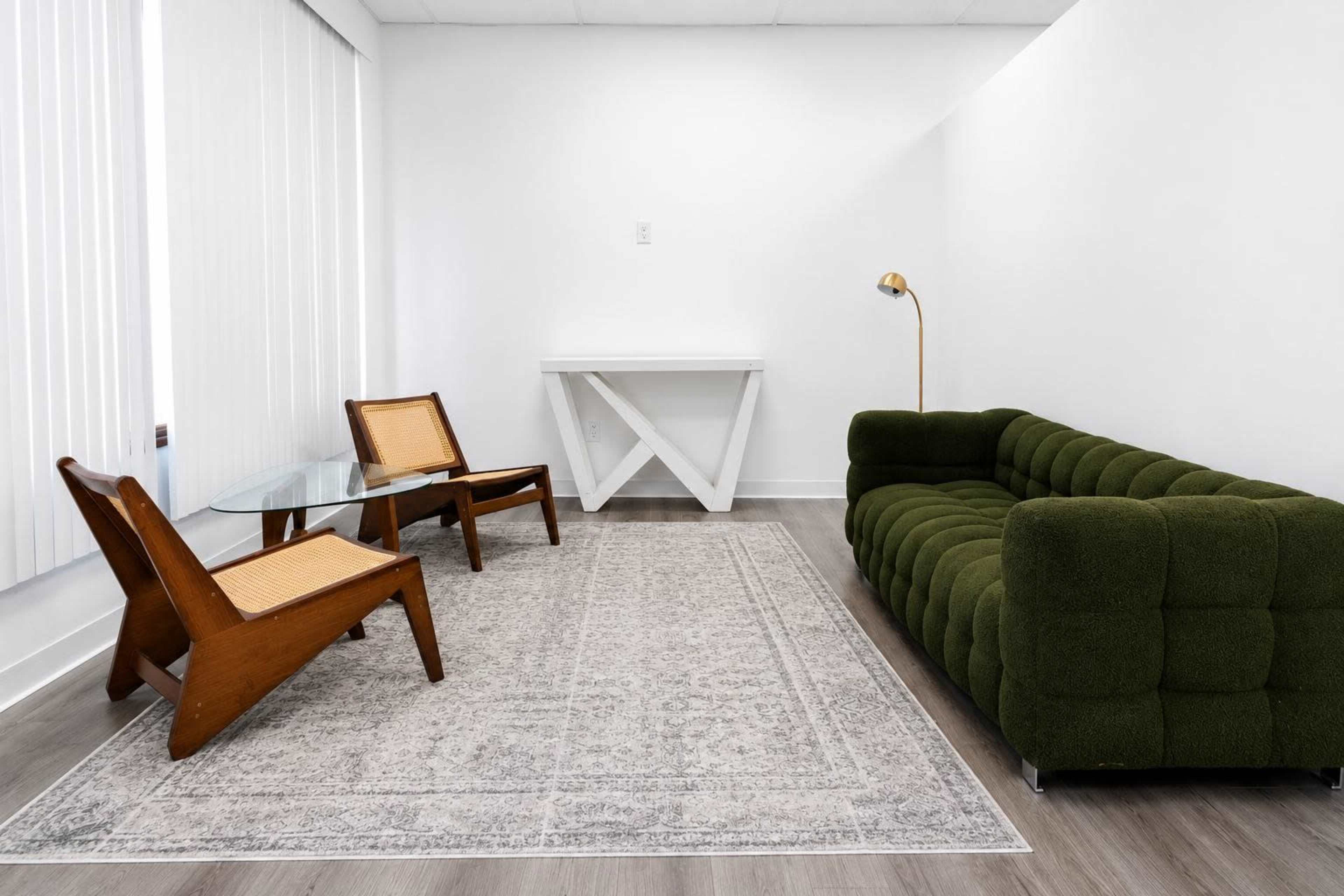The image shows a minimalist living room with a green tufted sofa, two wooden chairs, a glass table, and a patterned rug on a light wooden floor.