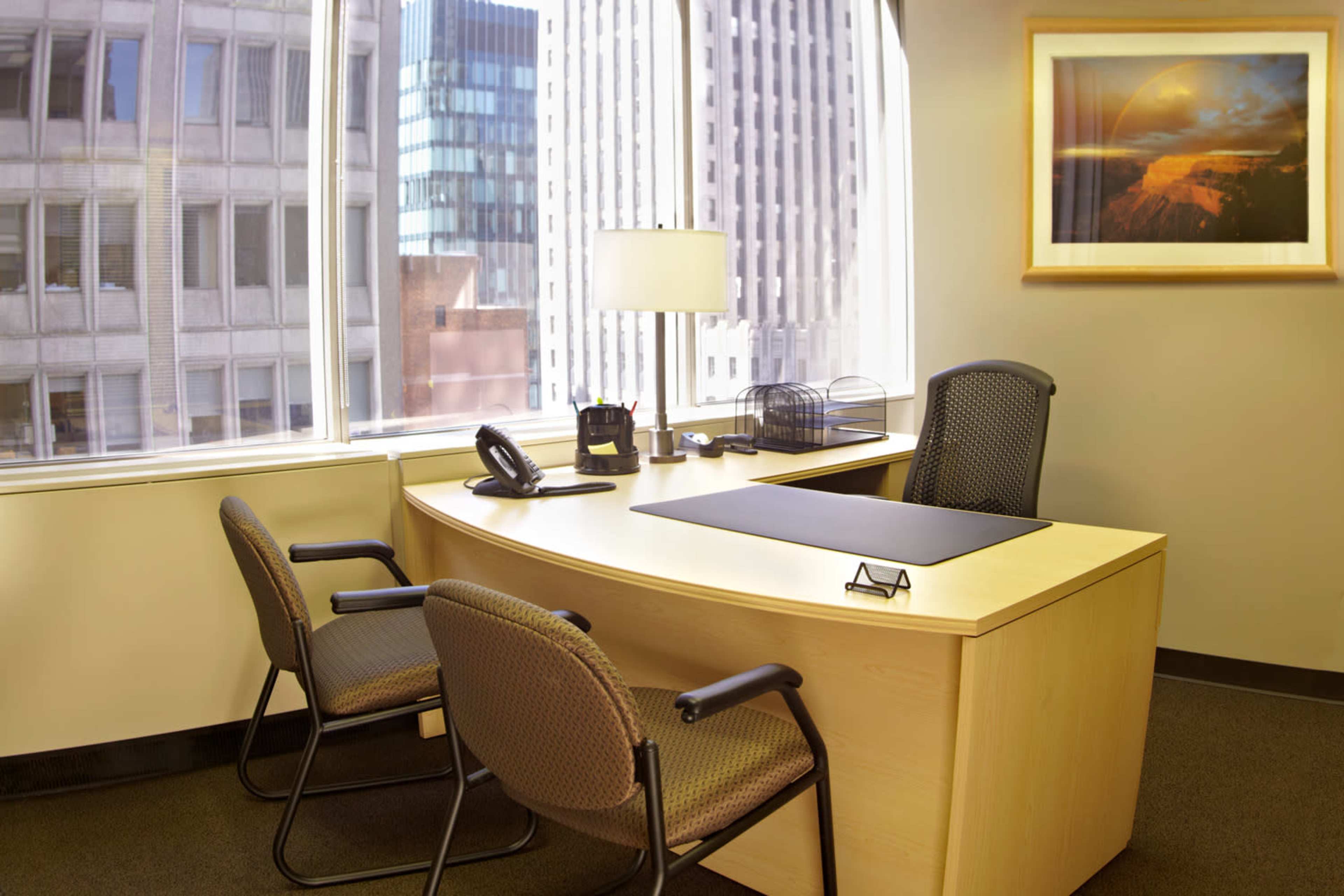 A modern office space featuring a curved wooden desk with two chairs, a desk lamp, and large windows overlooking a cityscape.