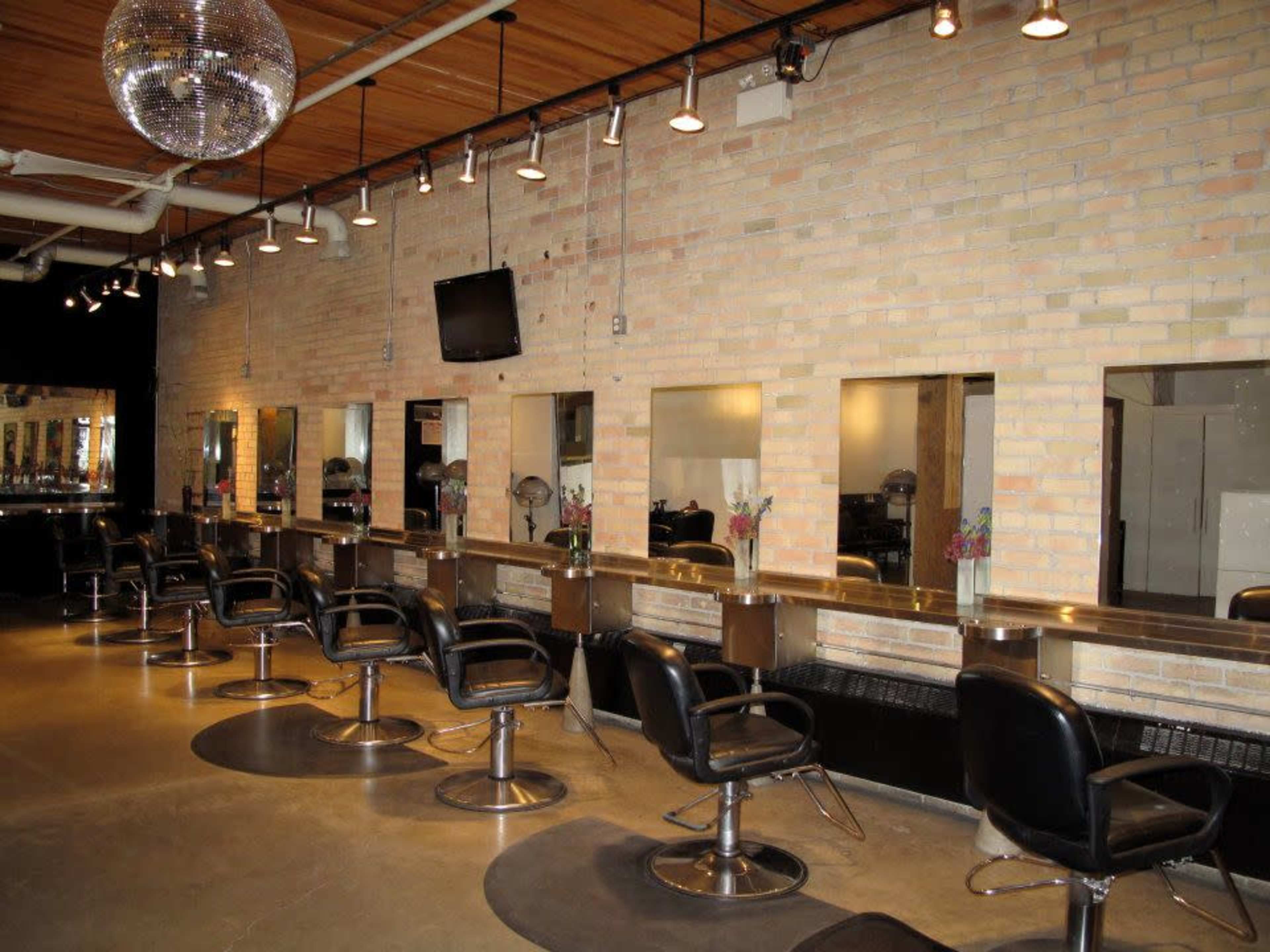 A modern salon interior features a row of styling stations with mirrors and adjustable chairs against a brick wall.