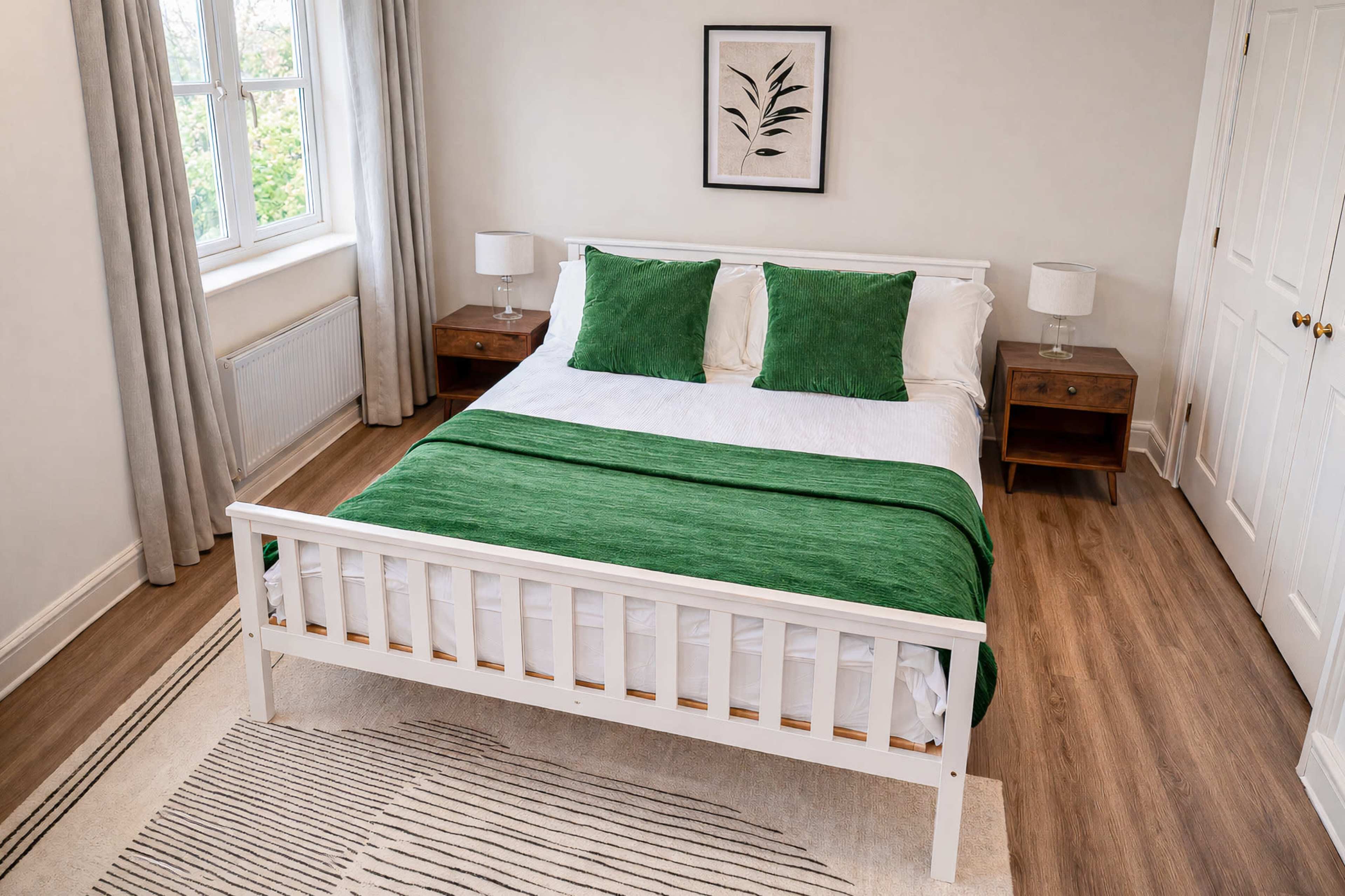 The image shows a neatly arranged bedroom with a white bed, green pillows and blanket, two bedside tables with lamps, and natural light coming through a window.