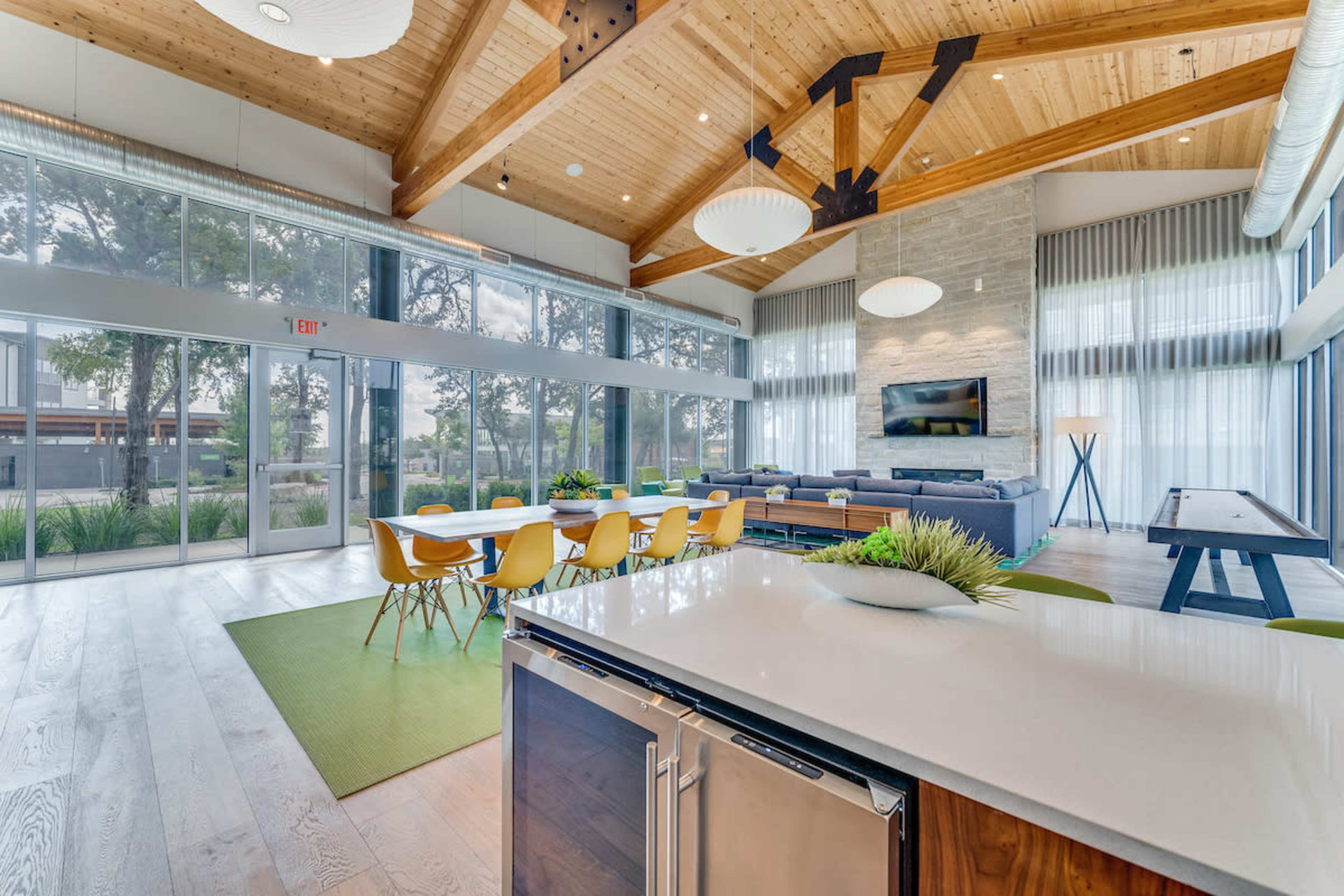 A spacious, modern interior with wooden beams, large windows, a dining area with yellow chairs, and a lounge area with a couch and TV.