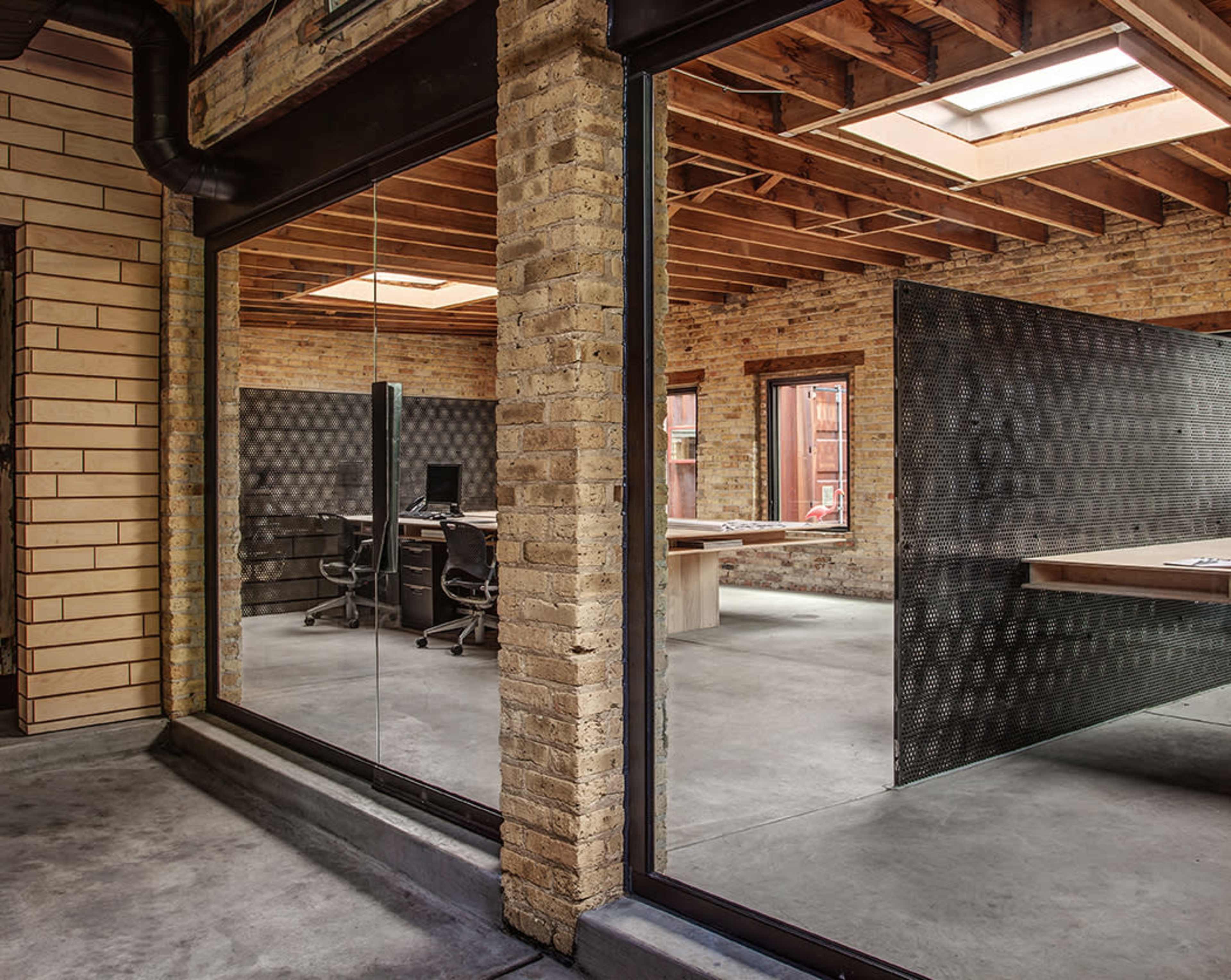 The image shows a modern office space with exposed brick walls and a large glass wall separating two work areas, one of which contains desks and chairs.