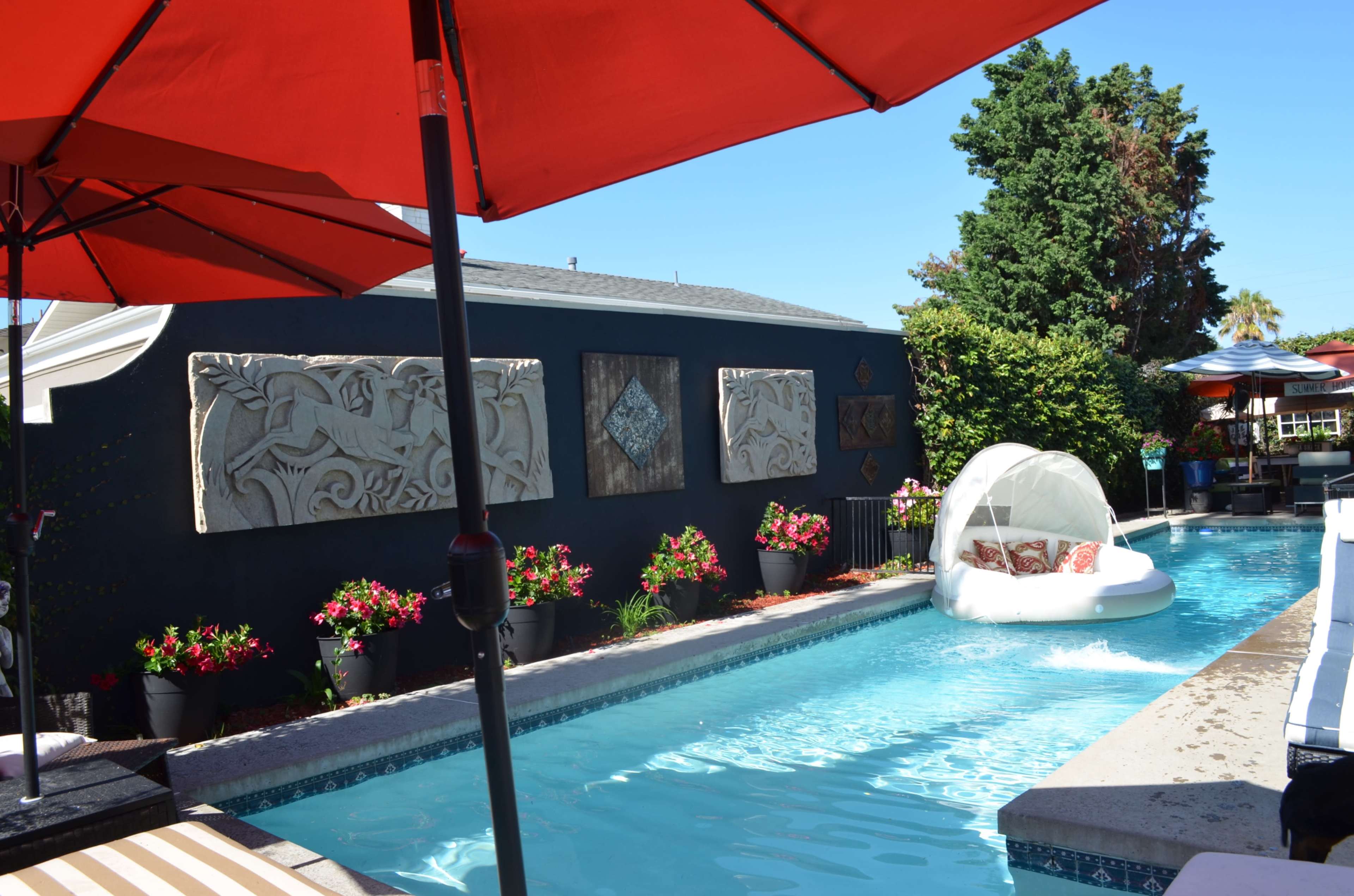 The image shows a pool area with a lounge chair and a white floating chair in the water, surrounded by potted plants and decorative wall panels.