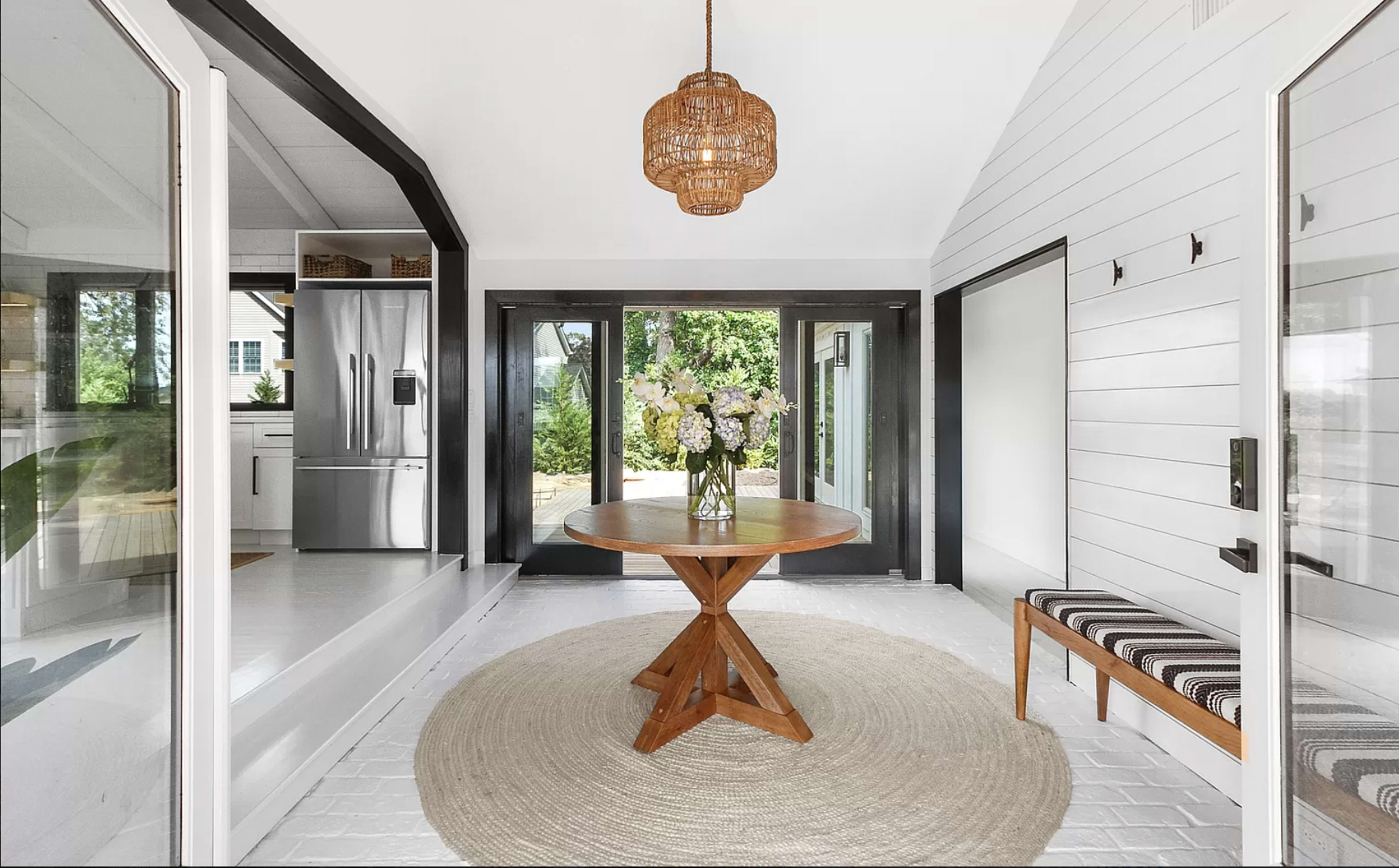 A bright entryway featuring a round wooden table, a woven pendant light, and a striped bench, with glass doors leading to a garden view.