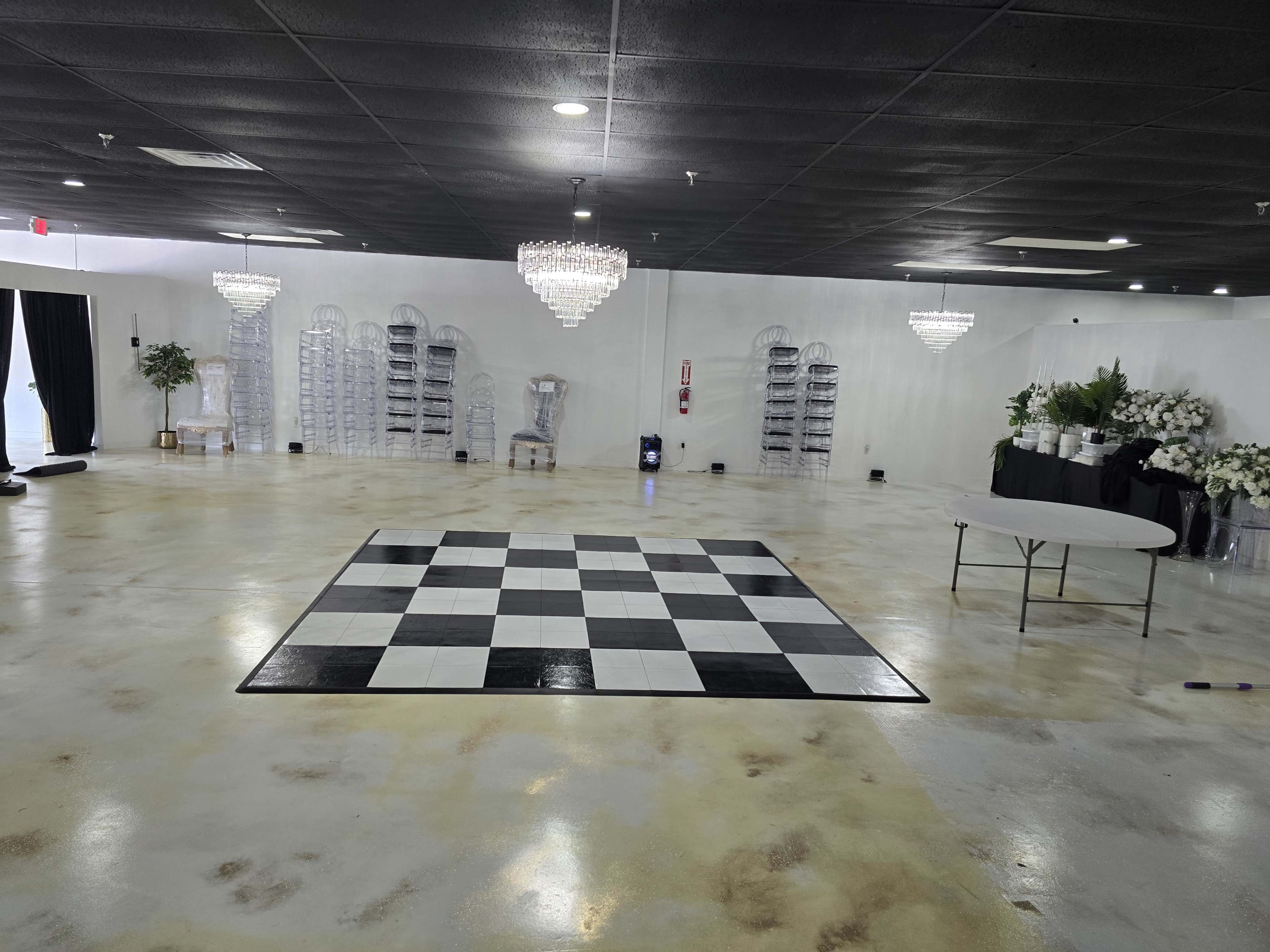 The image shows a spacious, empty event venue featuring a black and white checkered dance floor, two chandeliers, and several decorative arrangements along the walls.