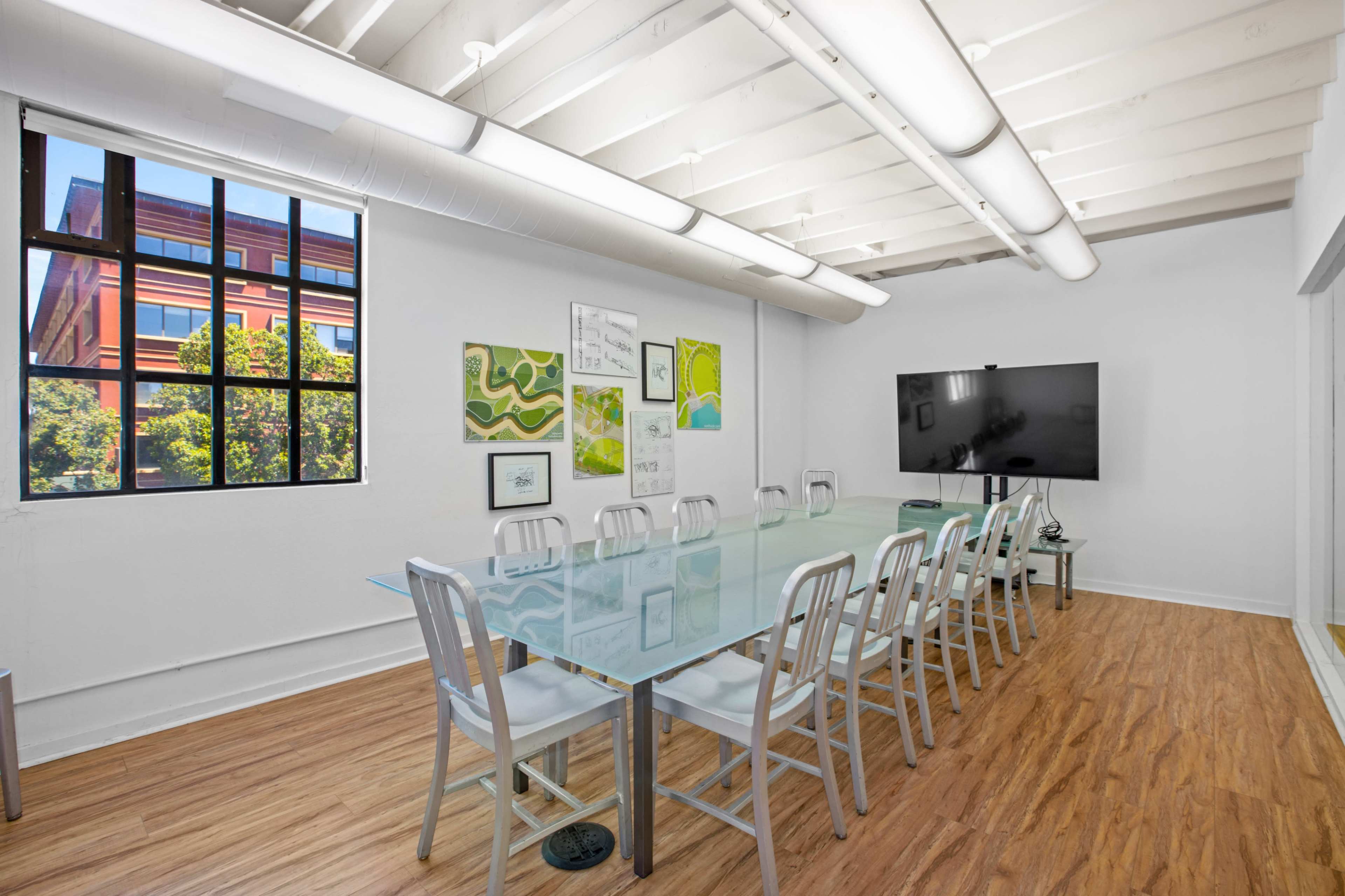 A conference room features a long glass table with multiple chairs, a large TV mounted on the wall, and large windows allowing natural light to enter.