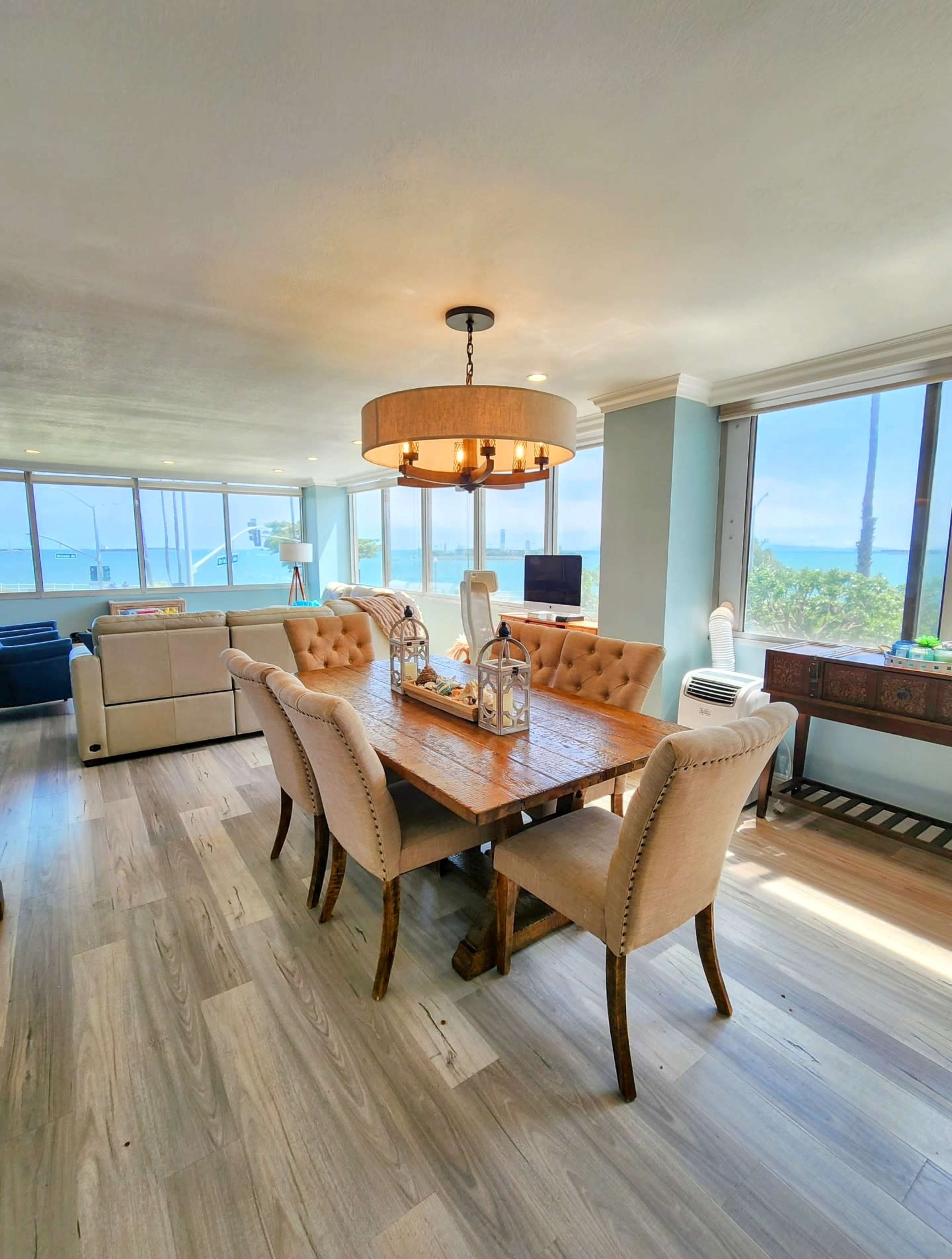 The image shows a dining area with a wooden table surrounded by upholstered chairs, illuminated by a large chandelier, and featuring windows that overlook a scenic view.
