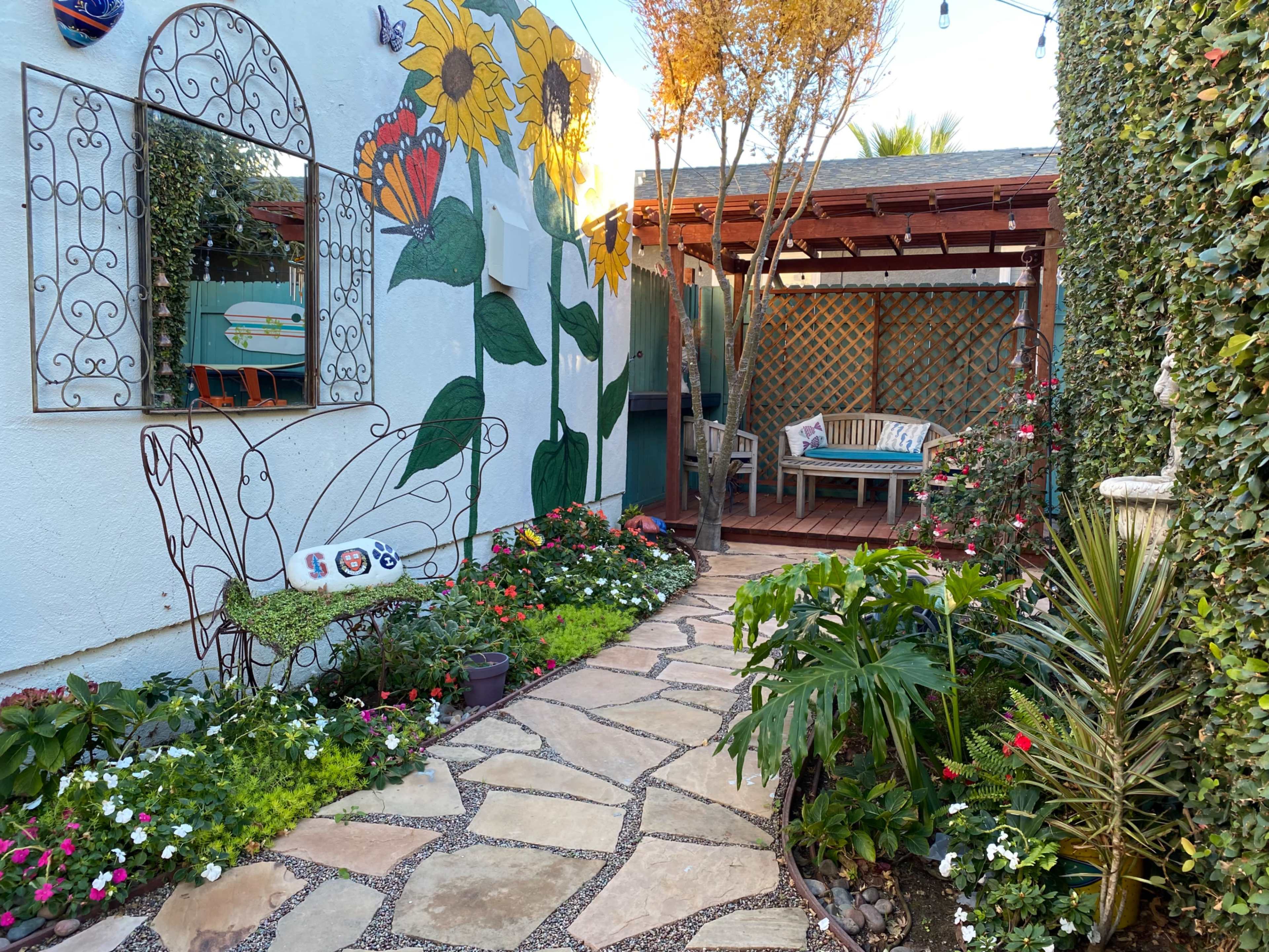 The image shows a garden pathway made of stone, leading to a seating area surrounded by colorful flowers and a mural of sunflowers on a wall.