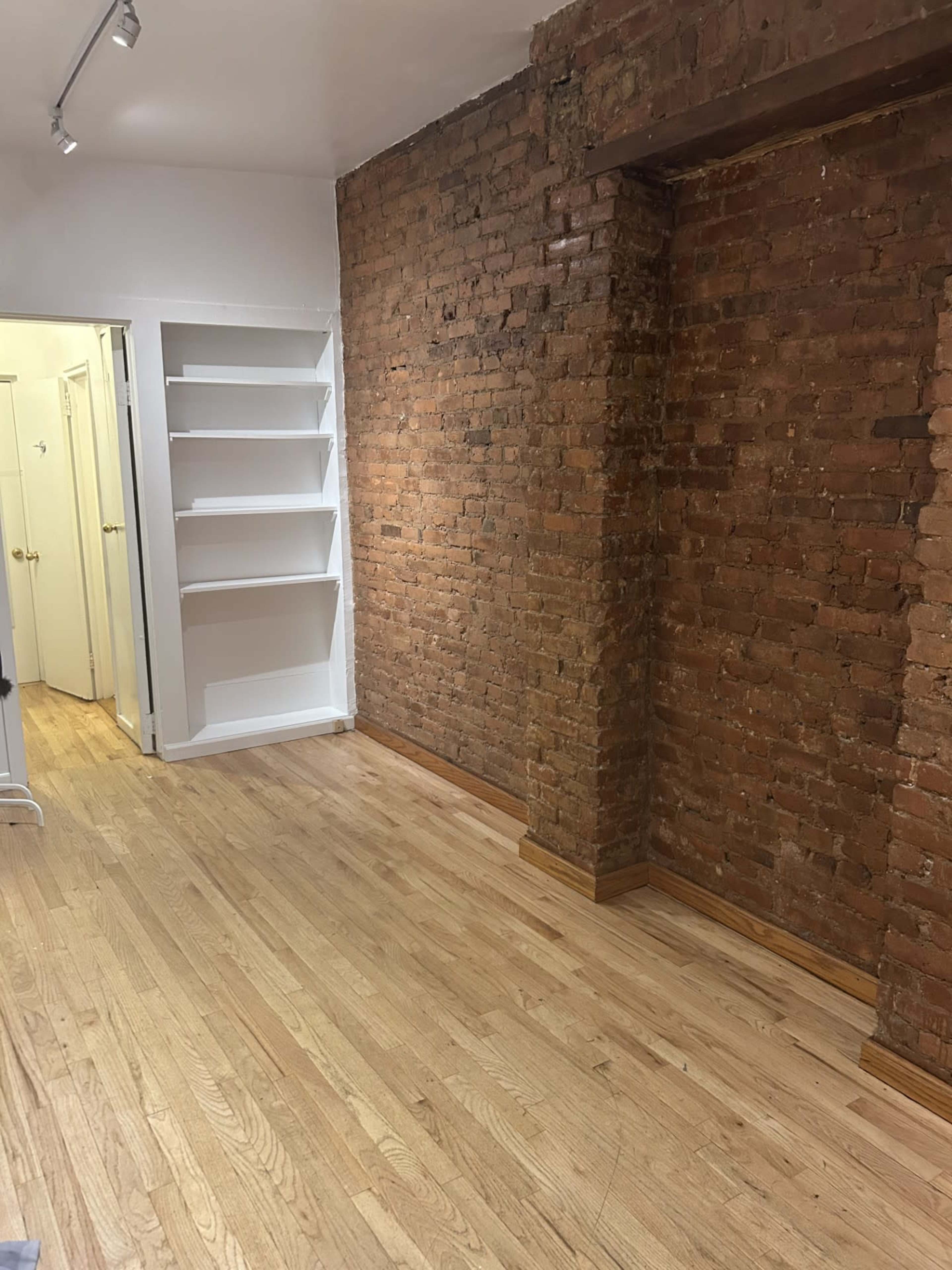 The image shows a room with exposed brick walls, wooden flooring, and a built-in shelf on one side.