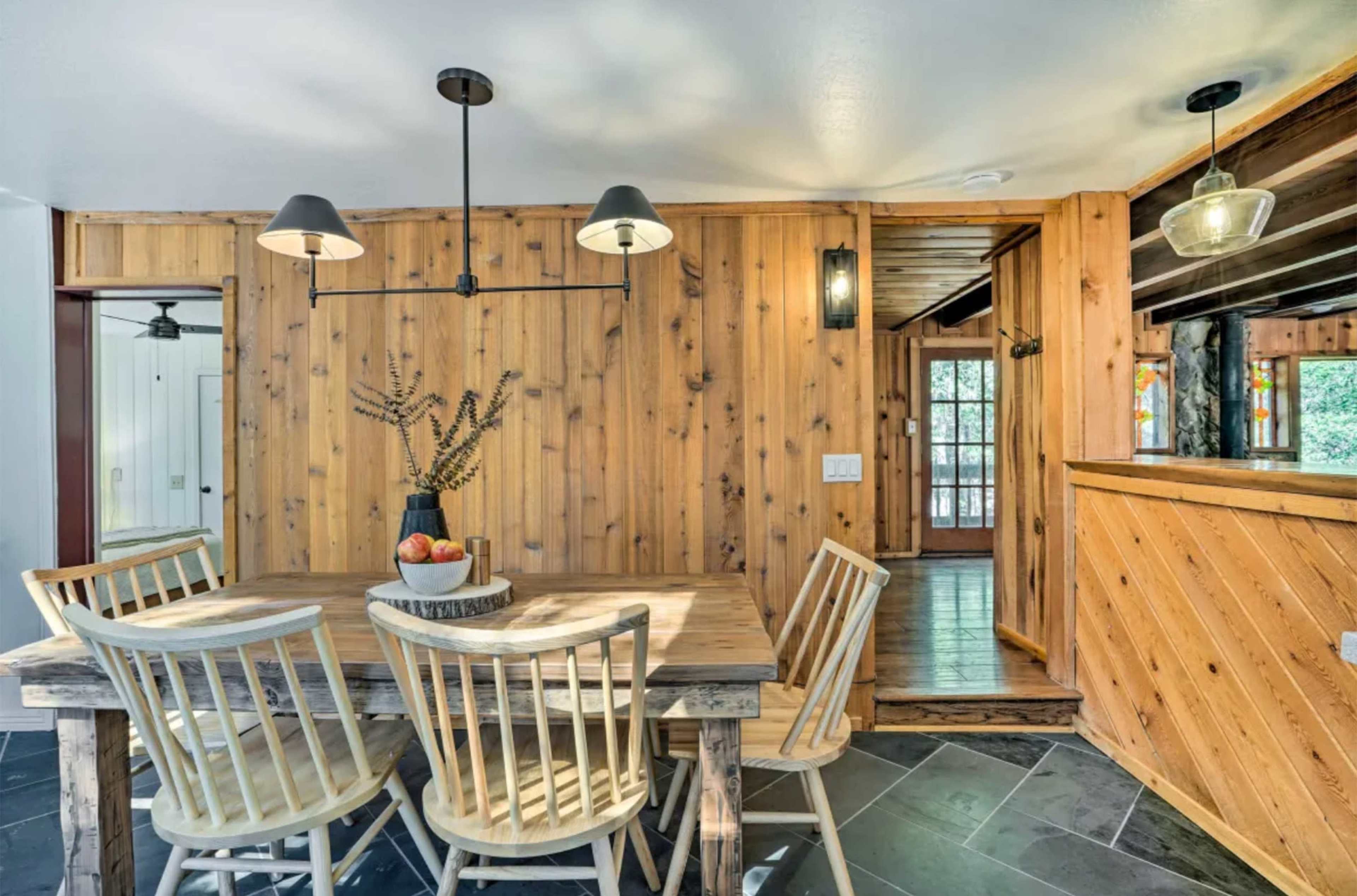 A wooden dining table with four chairs is positioned in a room featuring wooden walls and a door leading to a deck.