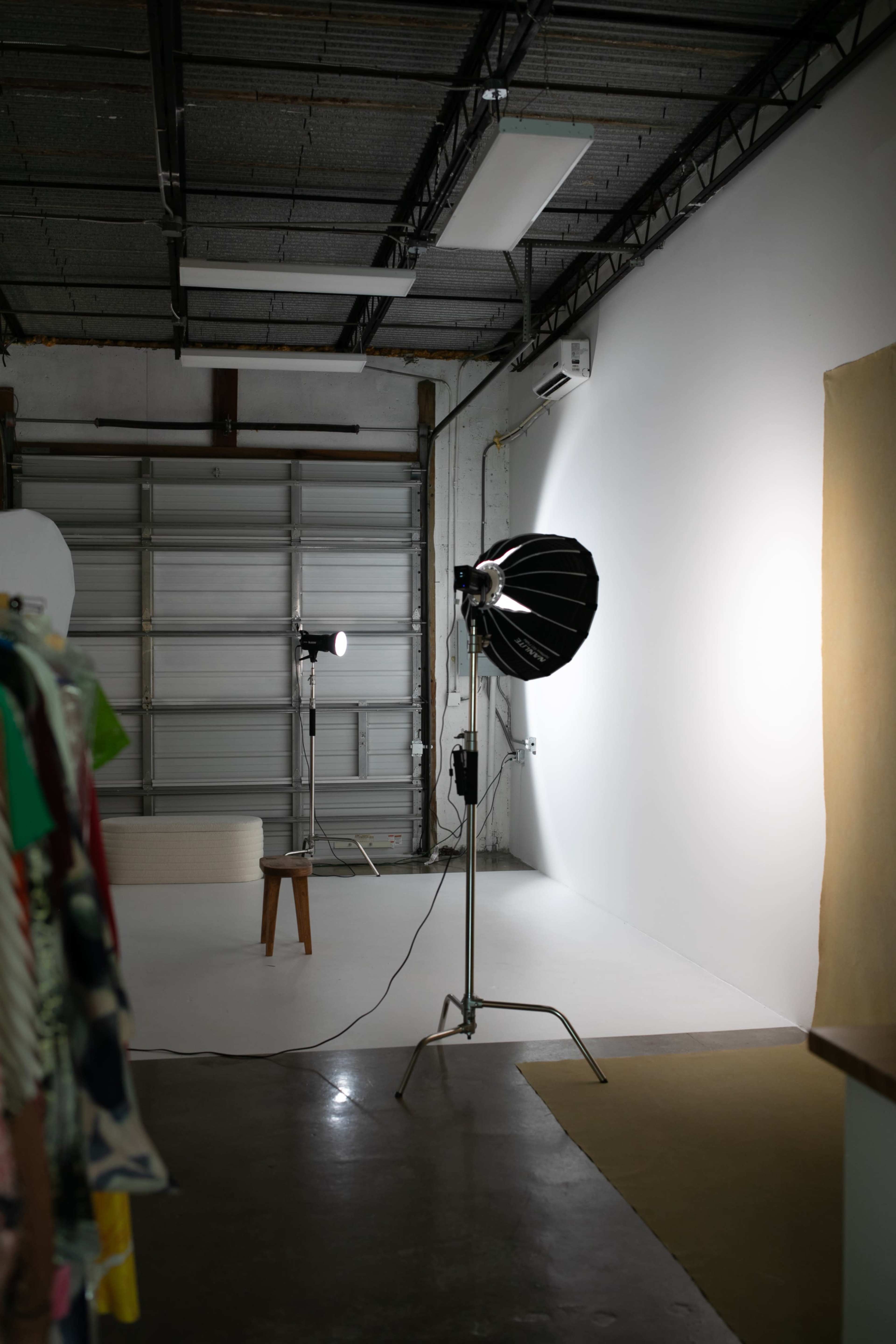 A photography studio features two light sources and a plain white wall, with clothing hanging on a rack in the foreground.