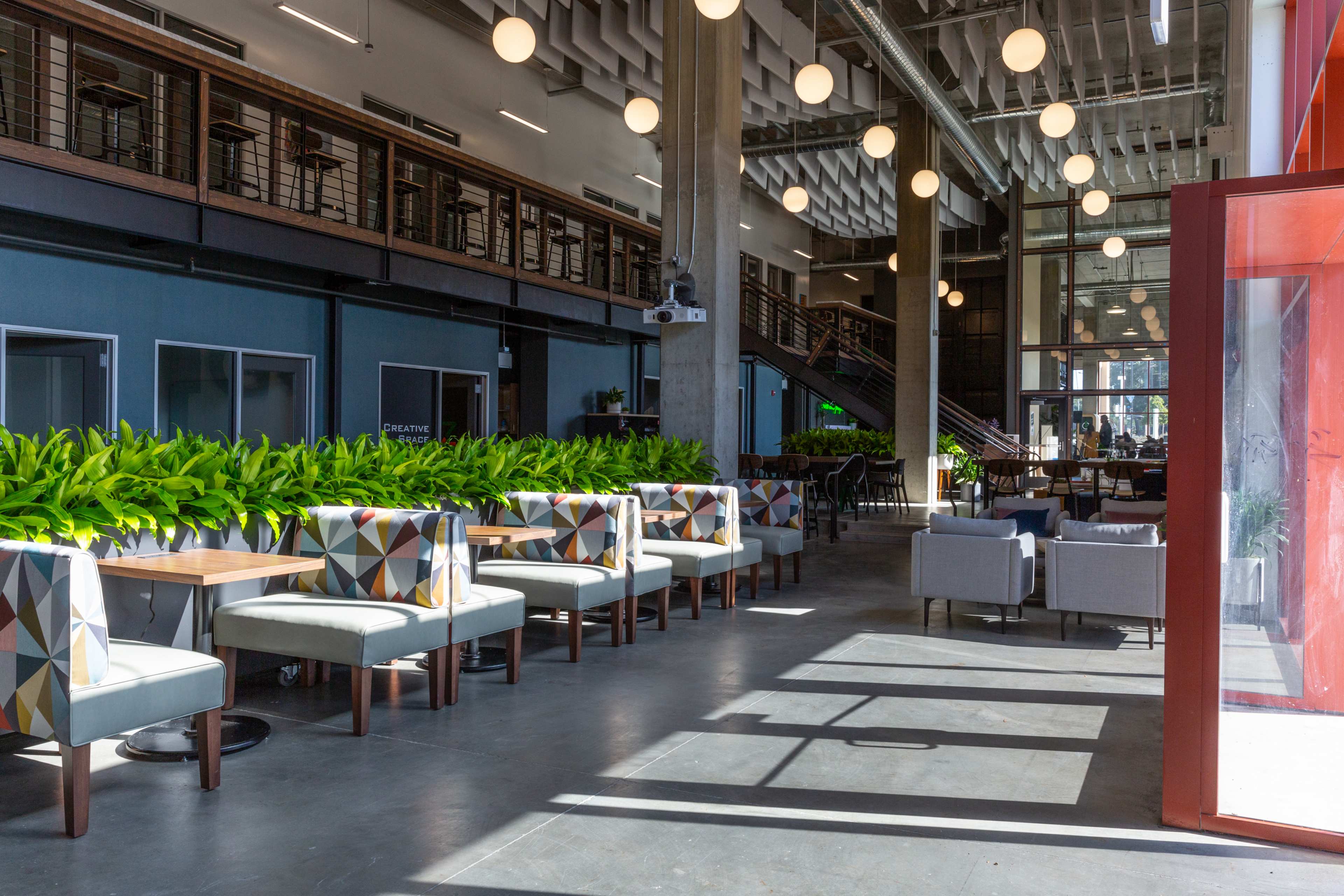 A modern indoor space with seating arrangements, large windows, and green plants, illuminated by hanging lights.