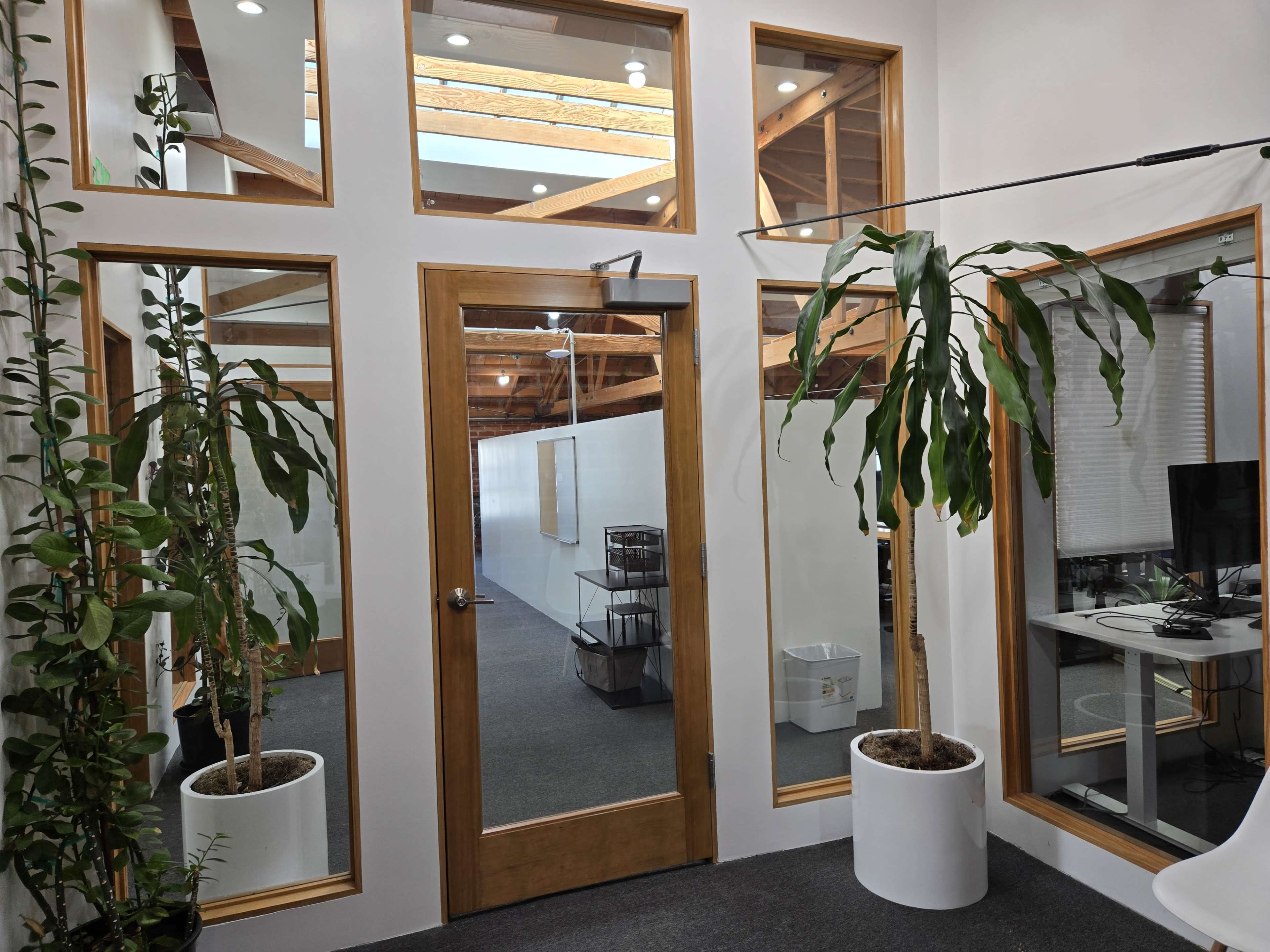 The image shows an interior office space featuring a glass door flanked by potted plants and large windows, with exposed wooden beams above.
