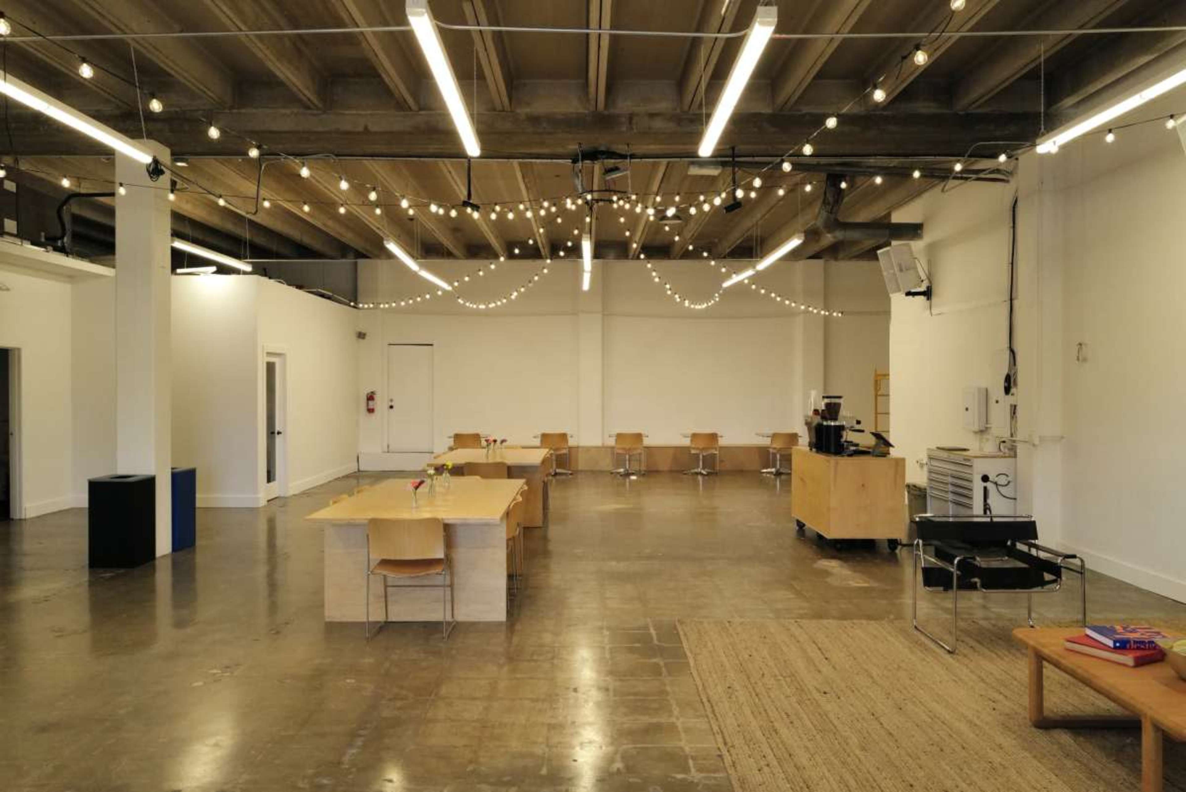 The image shows a spacious, well-lit room with wooden tables and chairs arranged for a communal setup, string lights hanging overhead, and a coffee station in one corner.