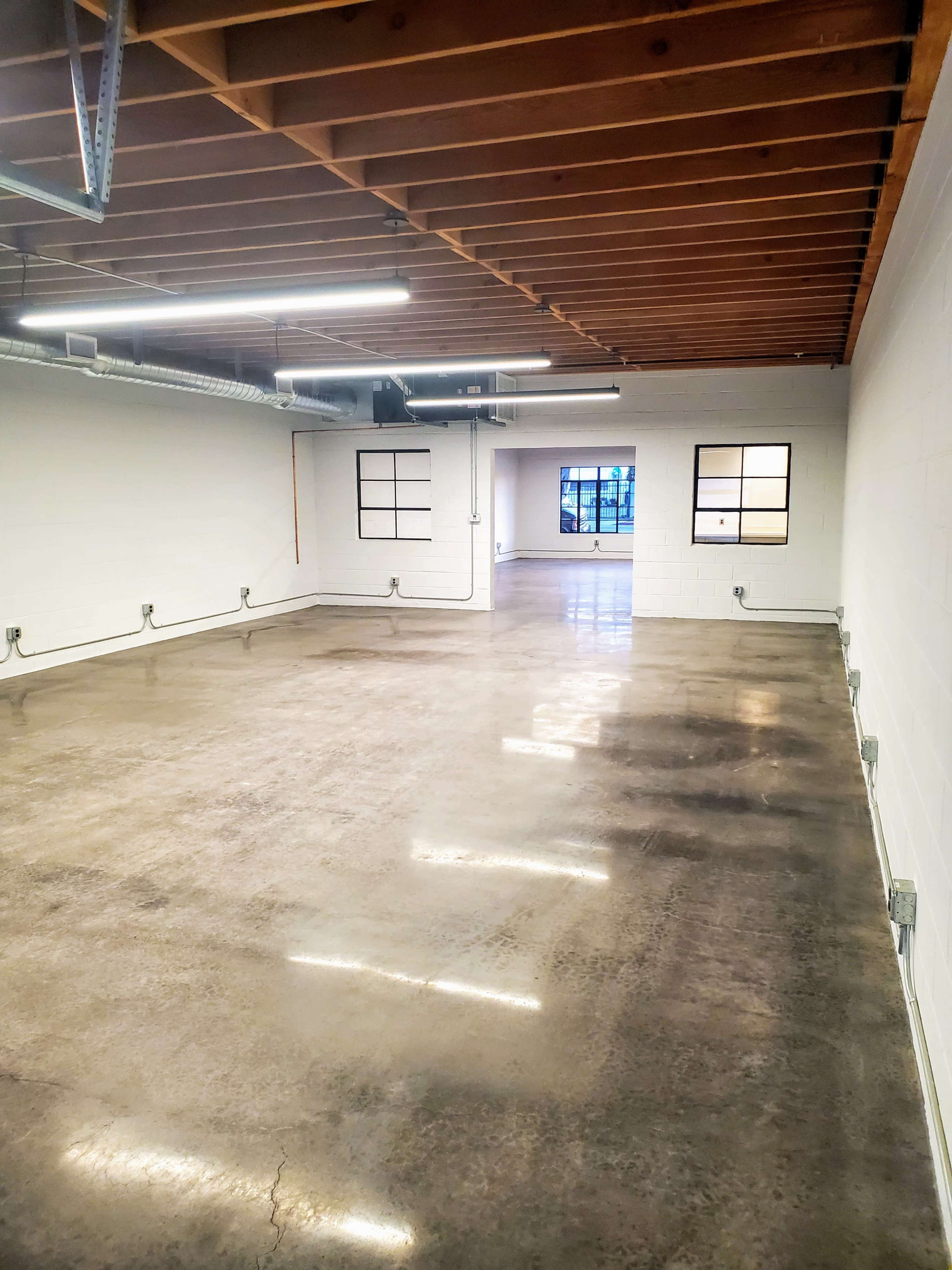 The image shows a spacious, minimally furnished room with a polished concrete floor, exposed wooden beams, and large windows at one end.