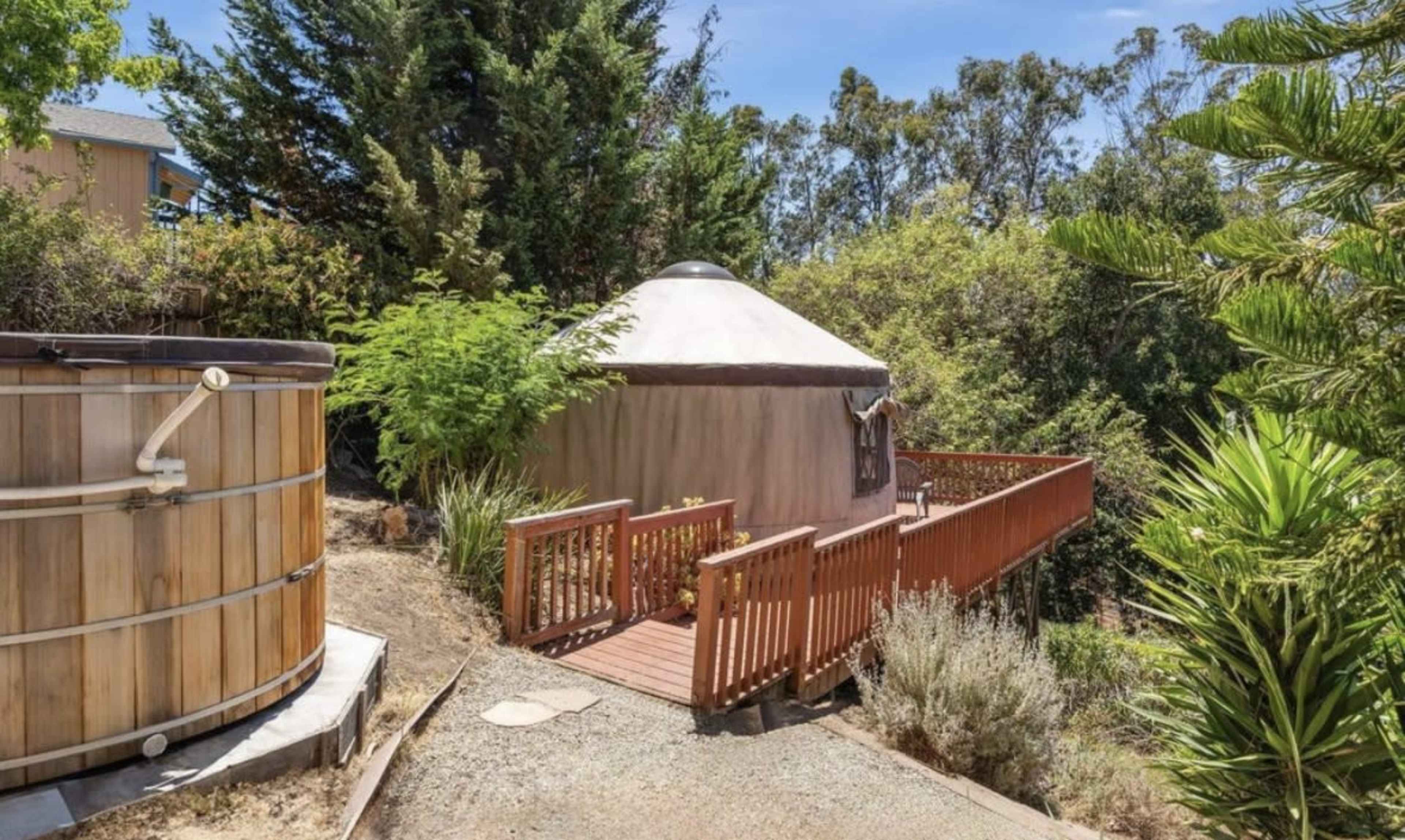 The image shows a wooden deck leading to a yurt surrounded by greenery, with a hot tub nearby.