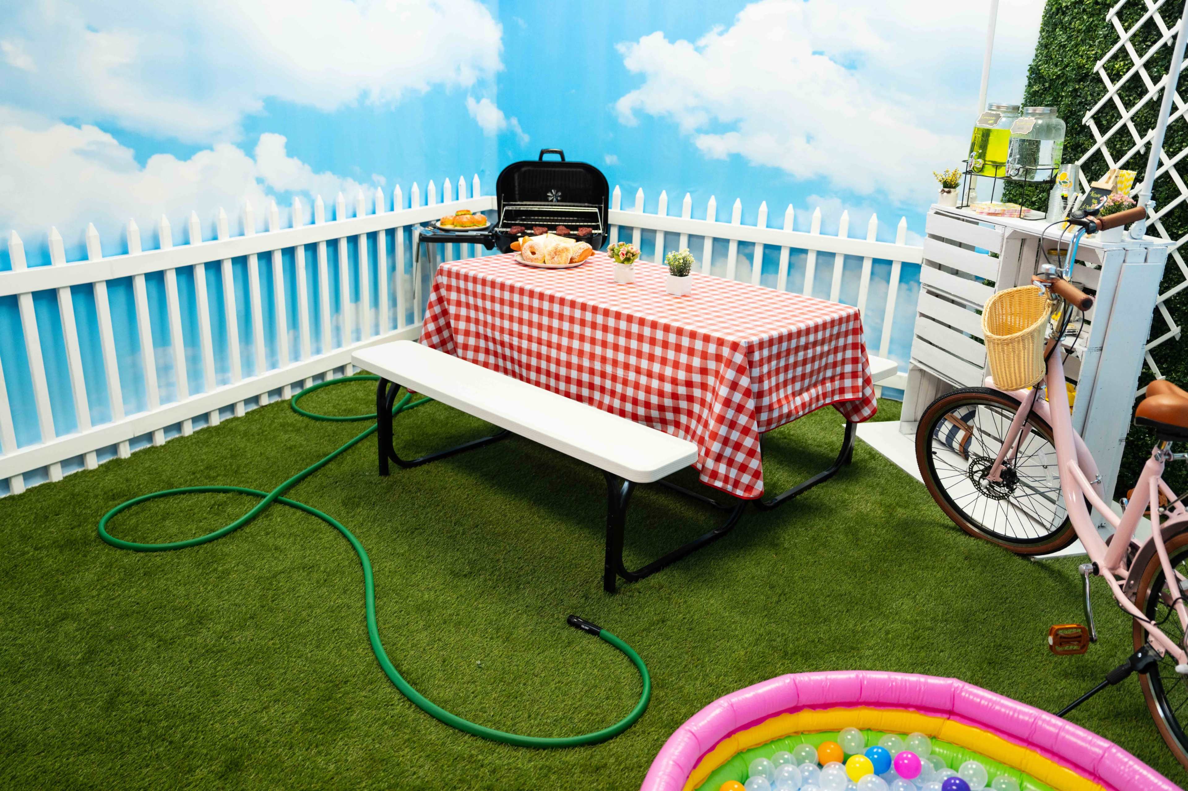 The scene features a picnic setup with a checkered tablecloth on a table, a grill, a hose, and a colorful inflatable pool in a fenced area adorned with a backdrop of clouds.