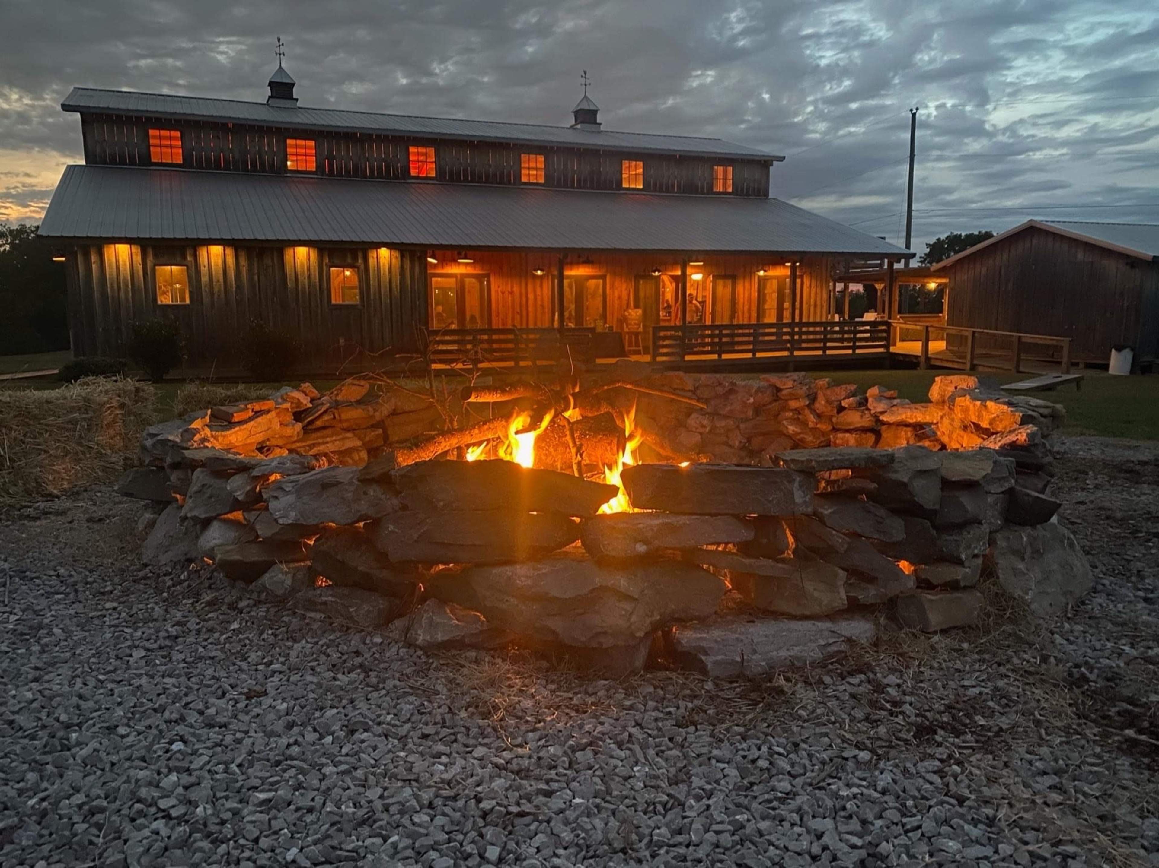 A stone fire pit filled with flames is situated in front of a wooden building with glowing windows under a cloudy sky.