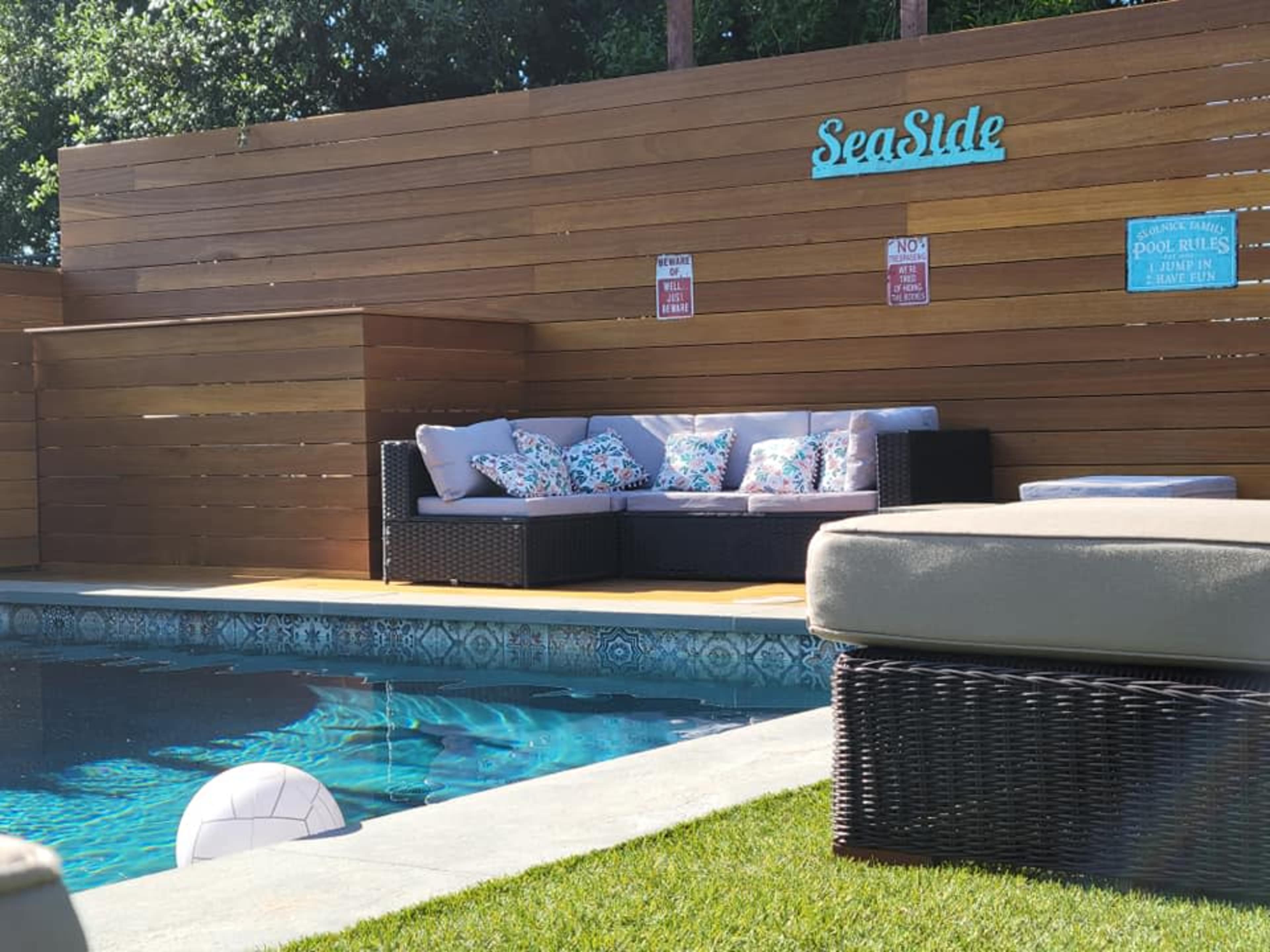 A swimming pool with a lounge area featuring a sectional sofa and decorative cushions is surrounded by wooden fencing and grass.