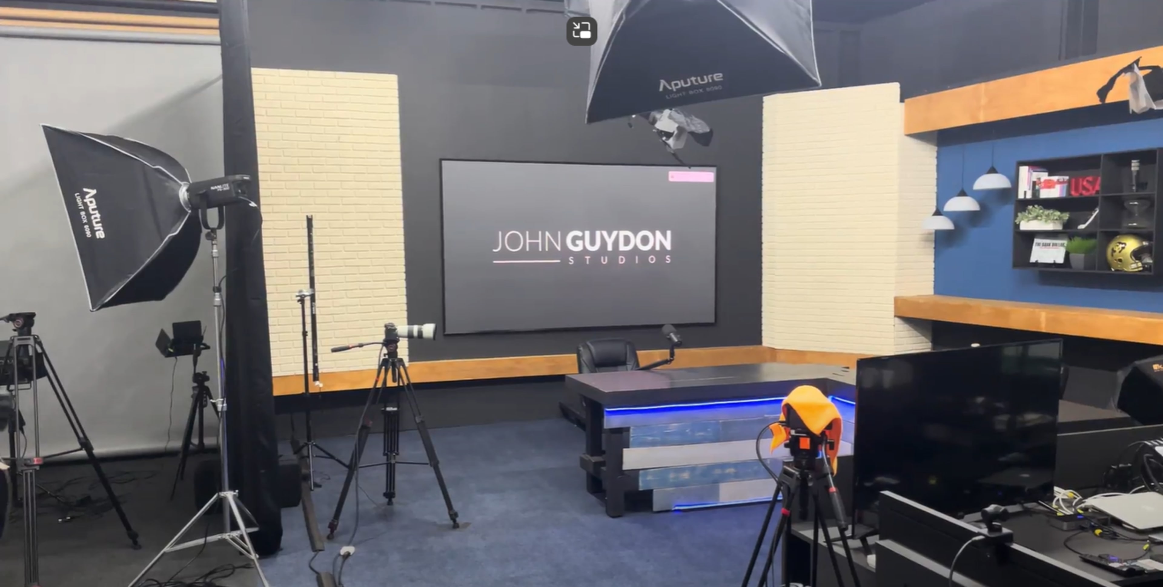 A video studio set up with cameras, lighting equipment, and a central desk featuring a backdrop that reads "JOHN GUYDON STUDIOS."