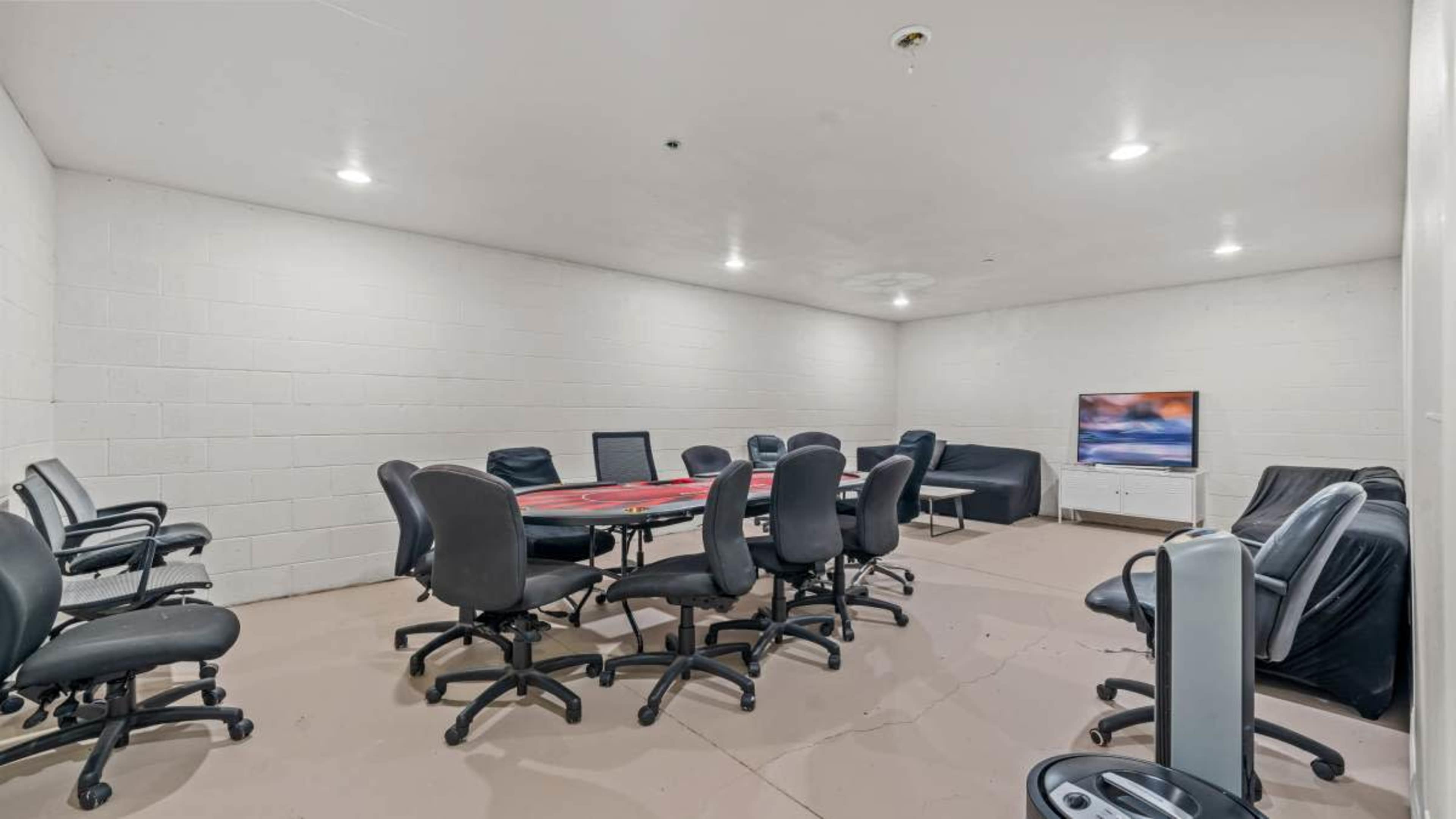 A basic meeting room features a round table surrounded by rolling chairs, with a television mounted on the wall.