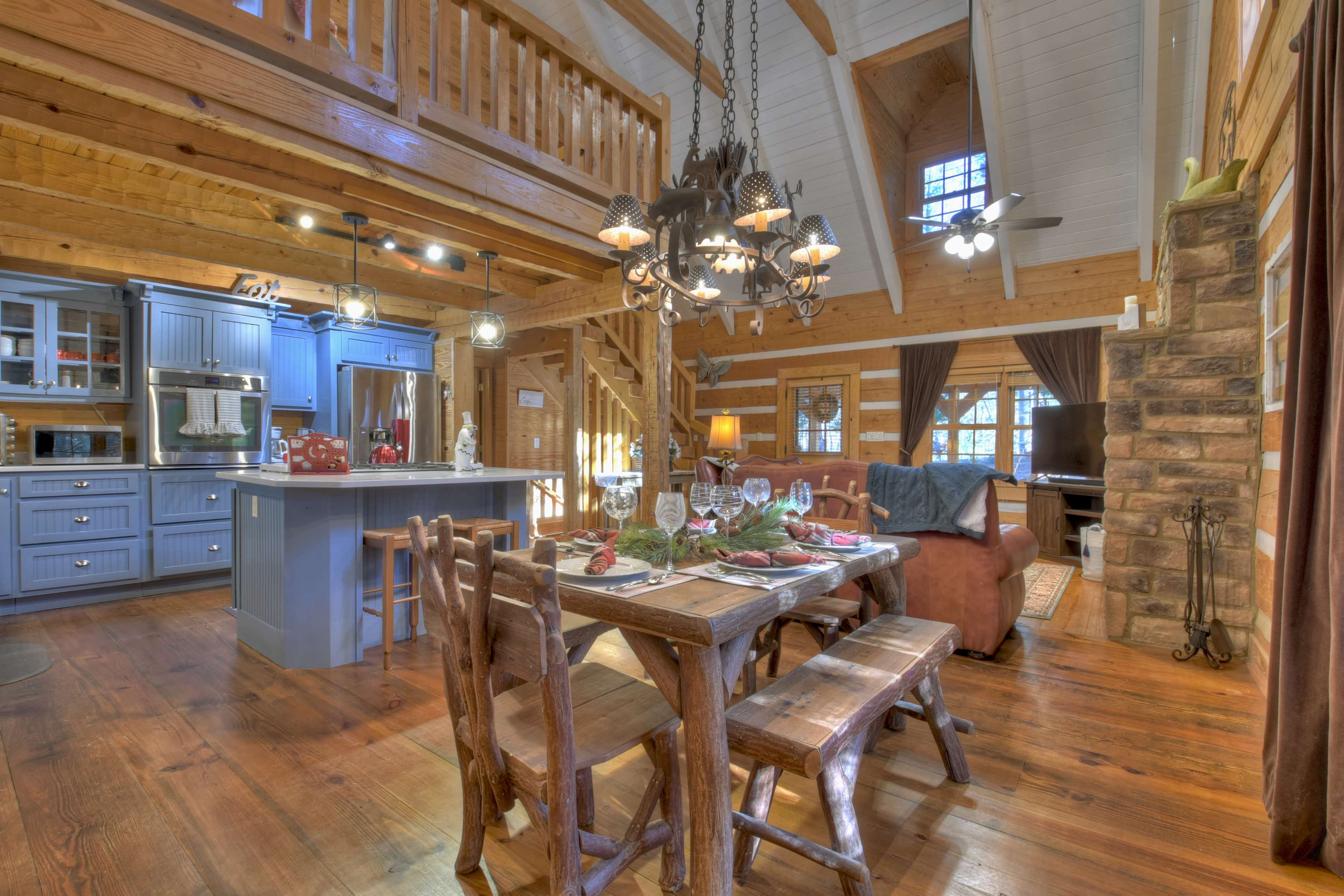 The interior of a rustic log cabin features a dining area with a wooden table, a kitchen with blue cabinetry, and a living area with a stone fireplace.
