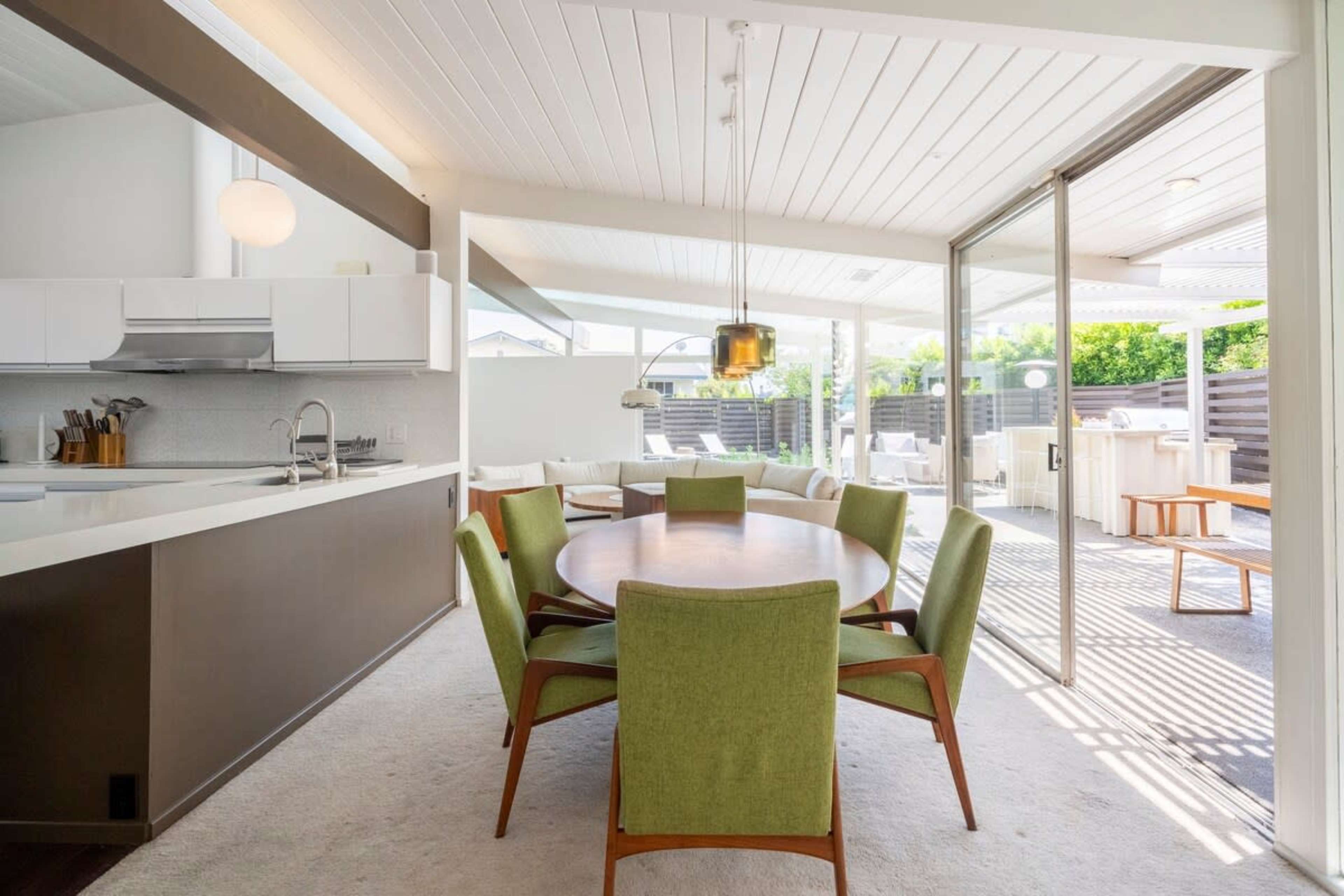 A modern dining area features a round wooden table surrounded by green chairs, with large windows opening to an outdoor space.