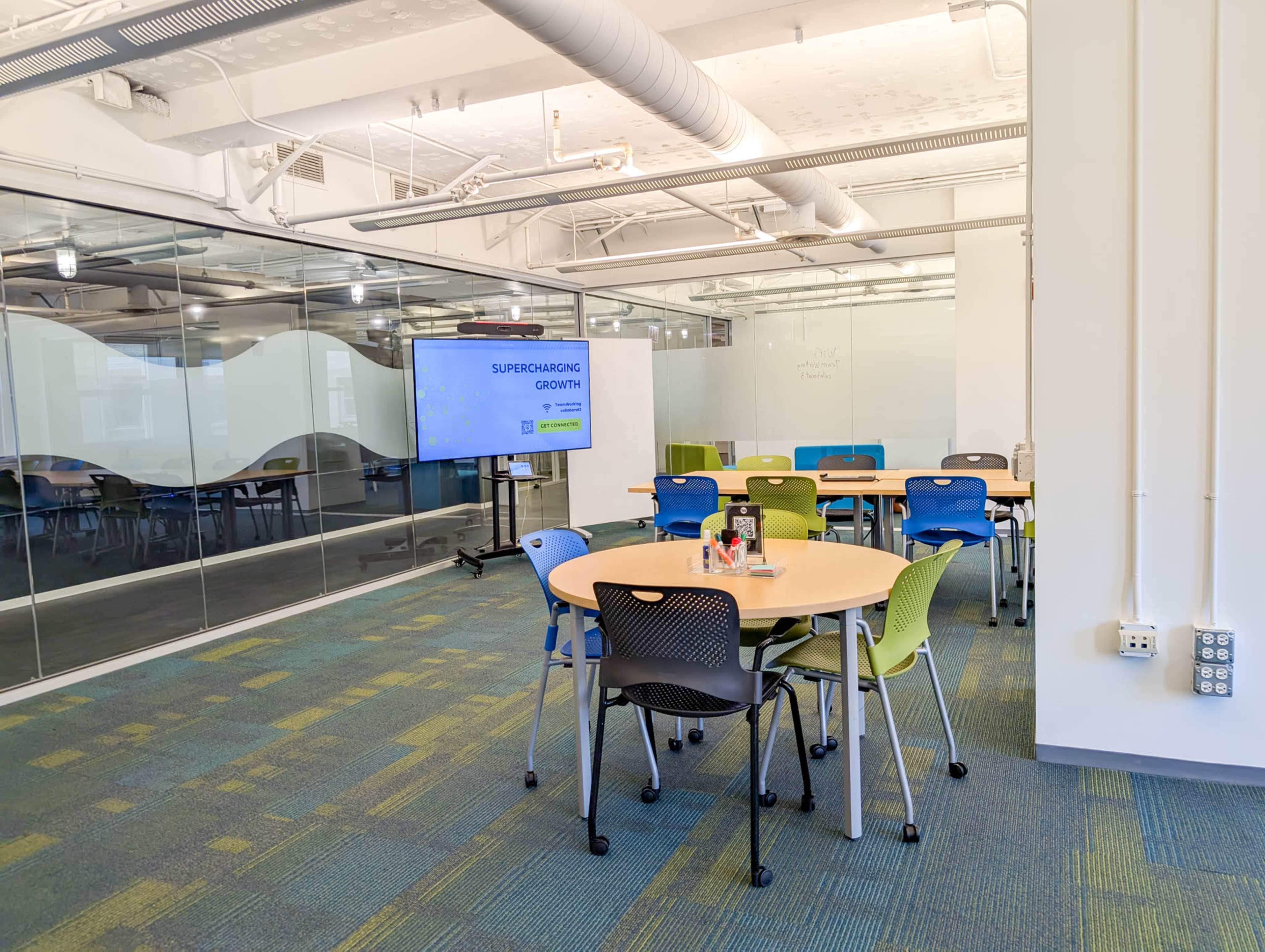 A modern meeting room features several round tables with colorful chairs, a large screen displaying a presentation, and glass walls separating it from adjacent spaces.