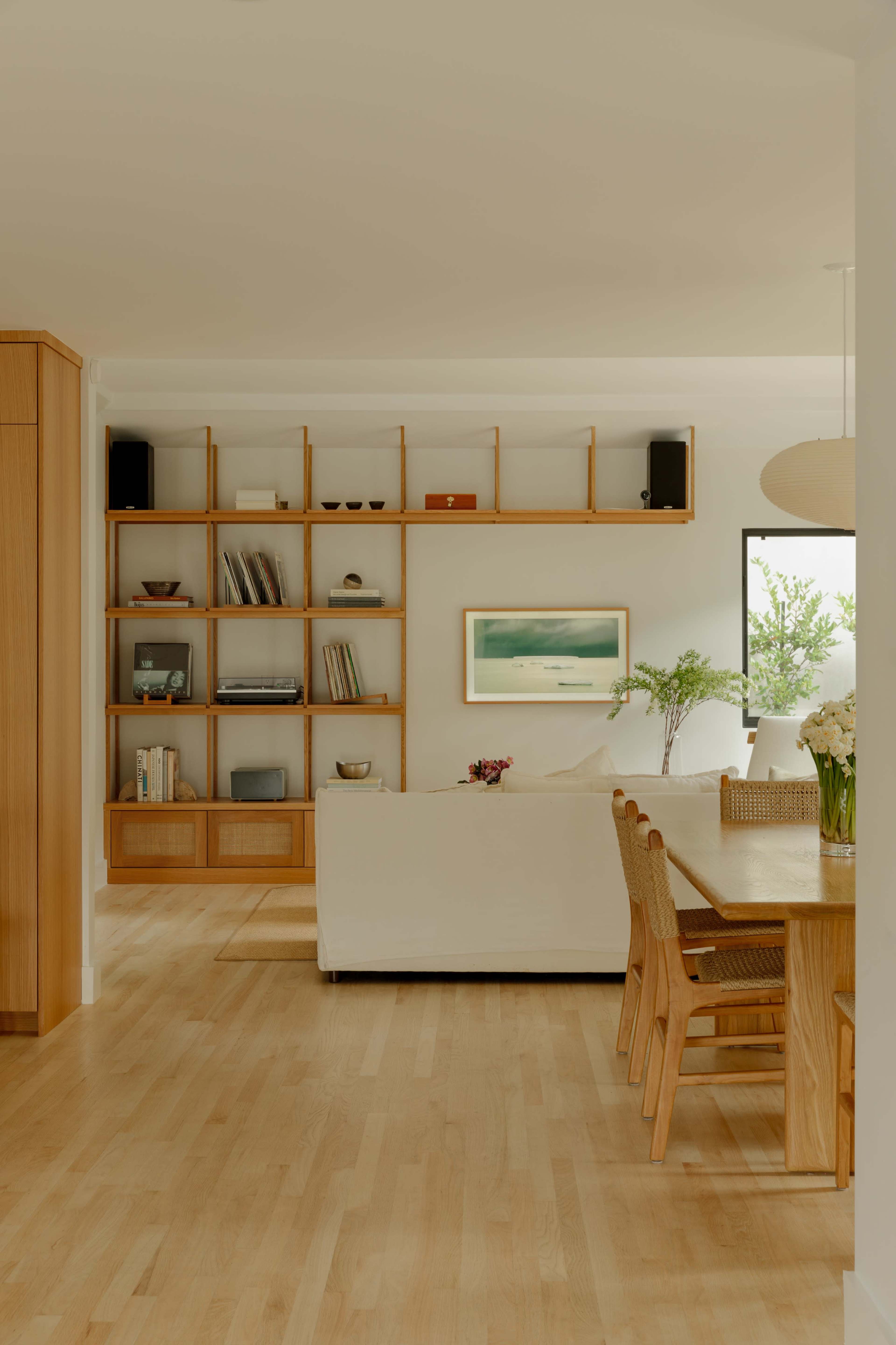 A bright, modern living space features a wooden shelving unit filled with books and decorative items, a white sofa, and a dining table surrounded by chairs.