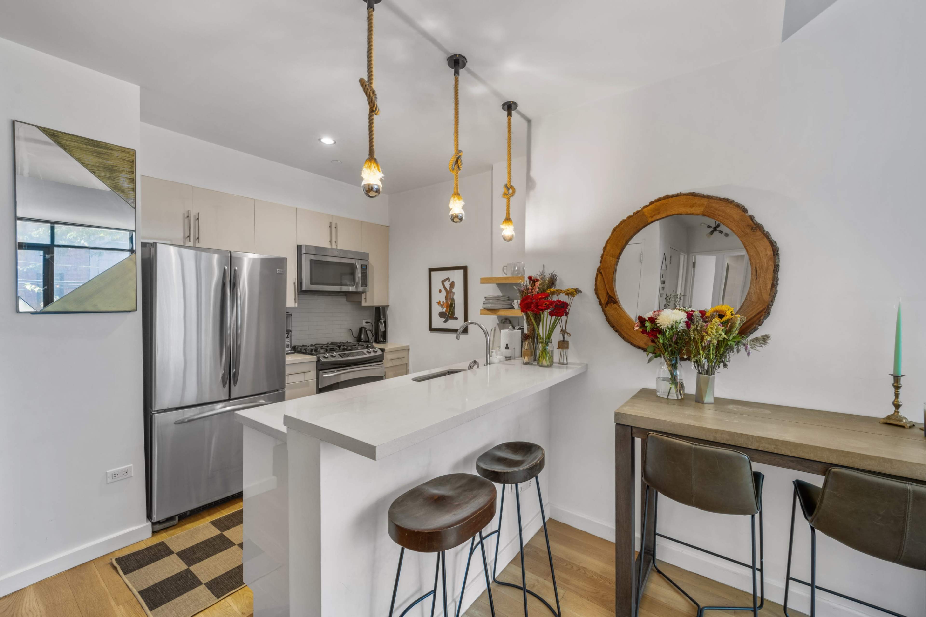 The image shows a modern kitchen with stainless steel appliances, a white countertop, and a bar with three stools, adorned with flowers and decorative elements.