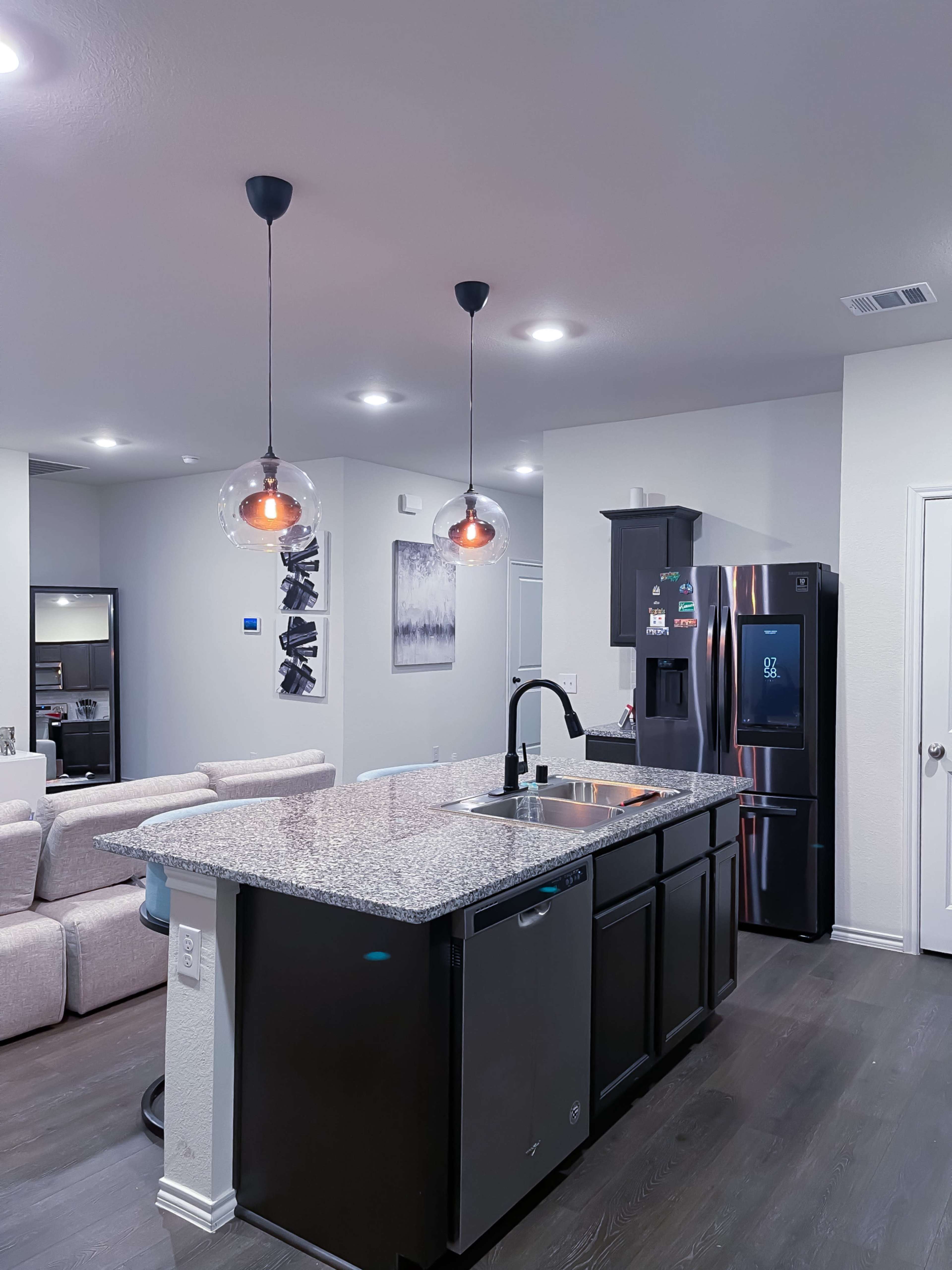 A modern kitchen features a large island with a sink and pendant lights, surrounded by a refrigerator and an open living area in the background.