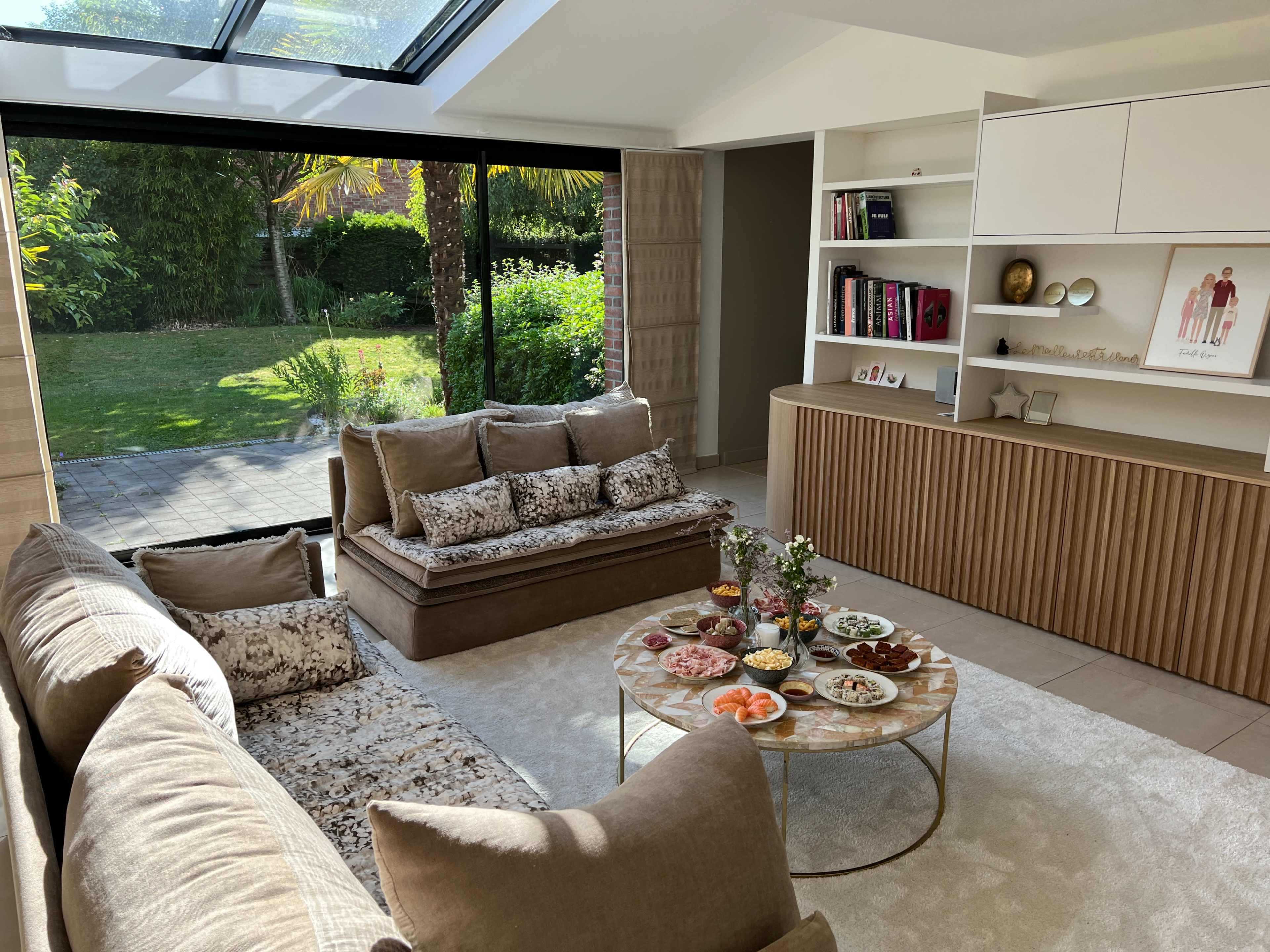 A well-lit living room features a comfortable seating arrangement with patterned cushions and a round coffee table displaying an array of food items, while large windows open to a view of a lush garden.